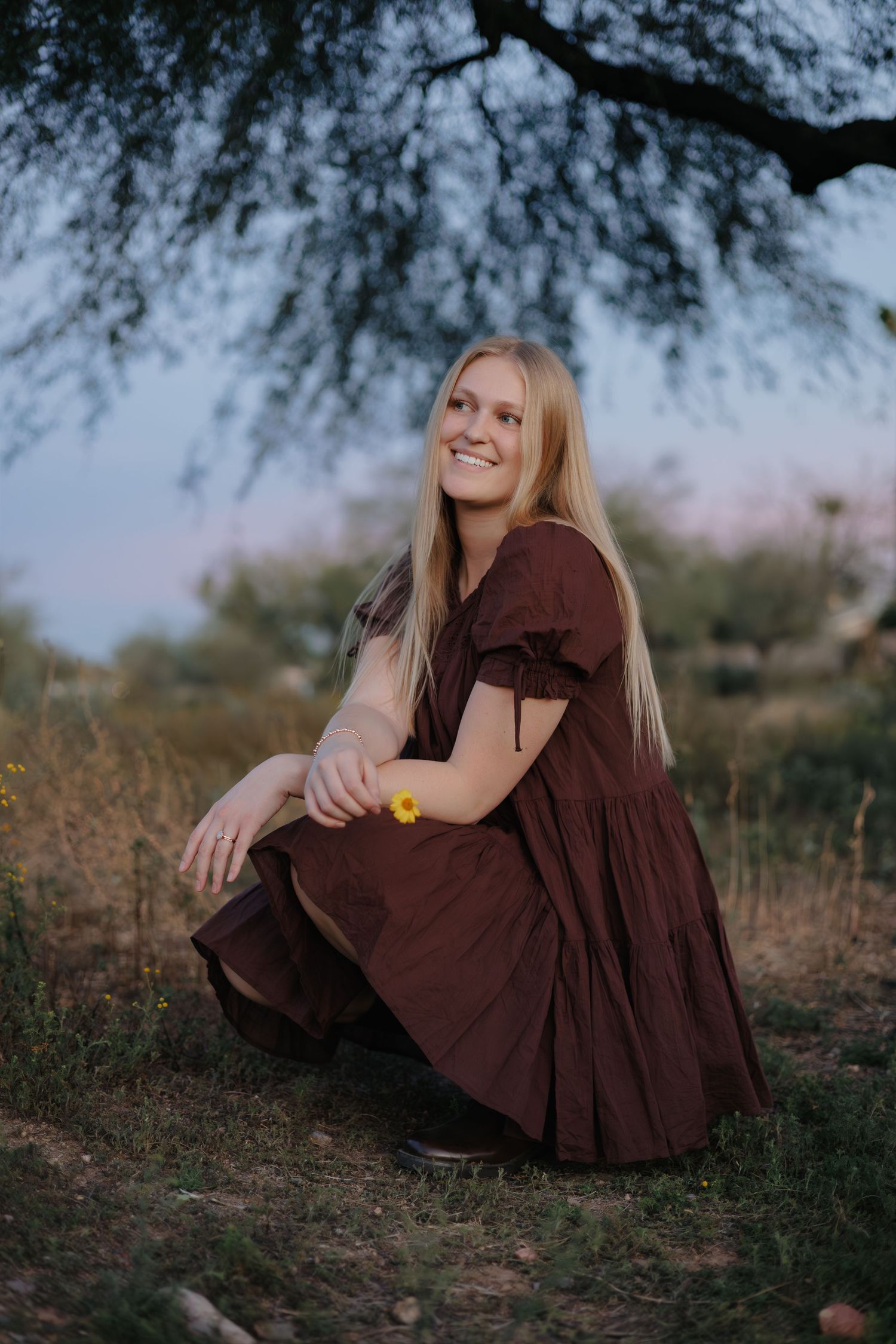 Person in brown dress sitting on desert ground under tree branches at sunset.