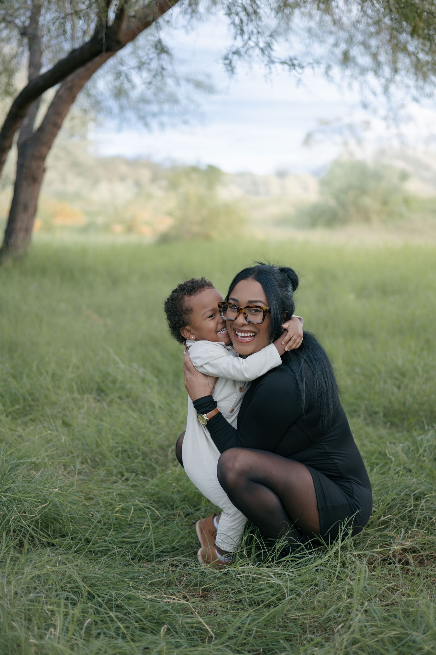 Mother and child sharing joyful embrace while lying in green grass under tree.