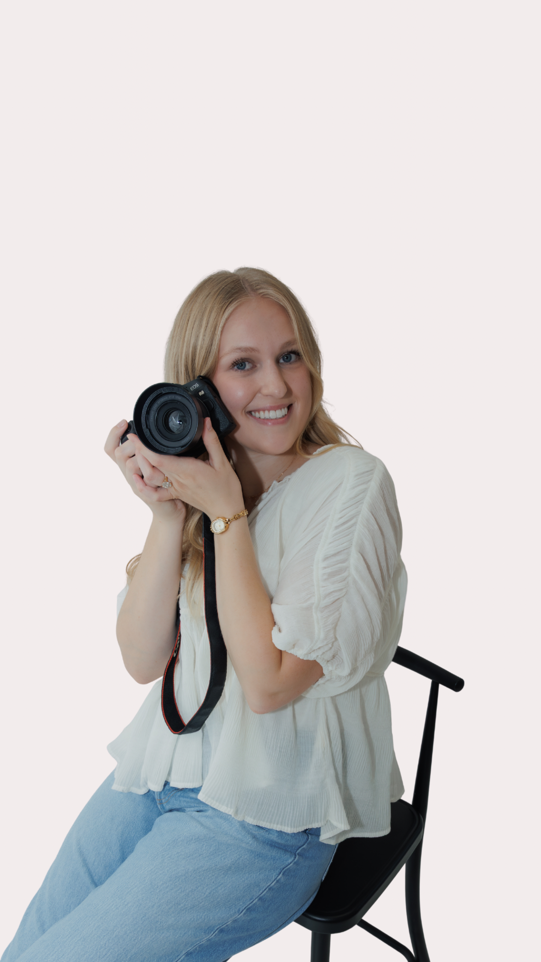 A photographer in light blue jeans and white top poses with a DSLR camera while sitting in a black chair.