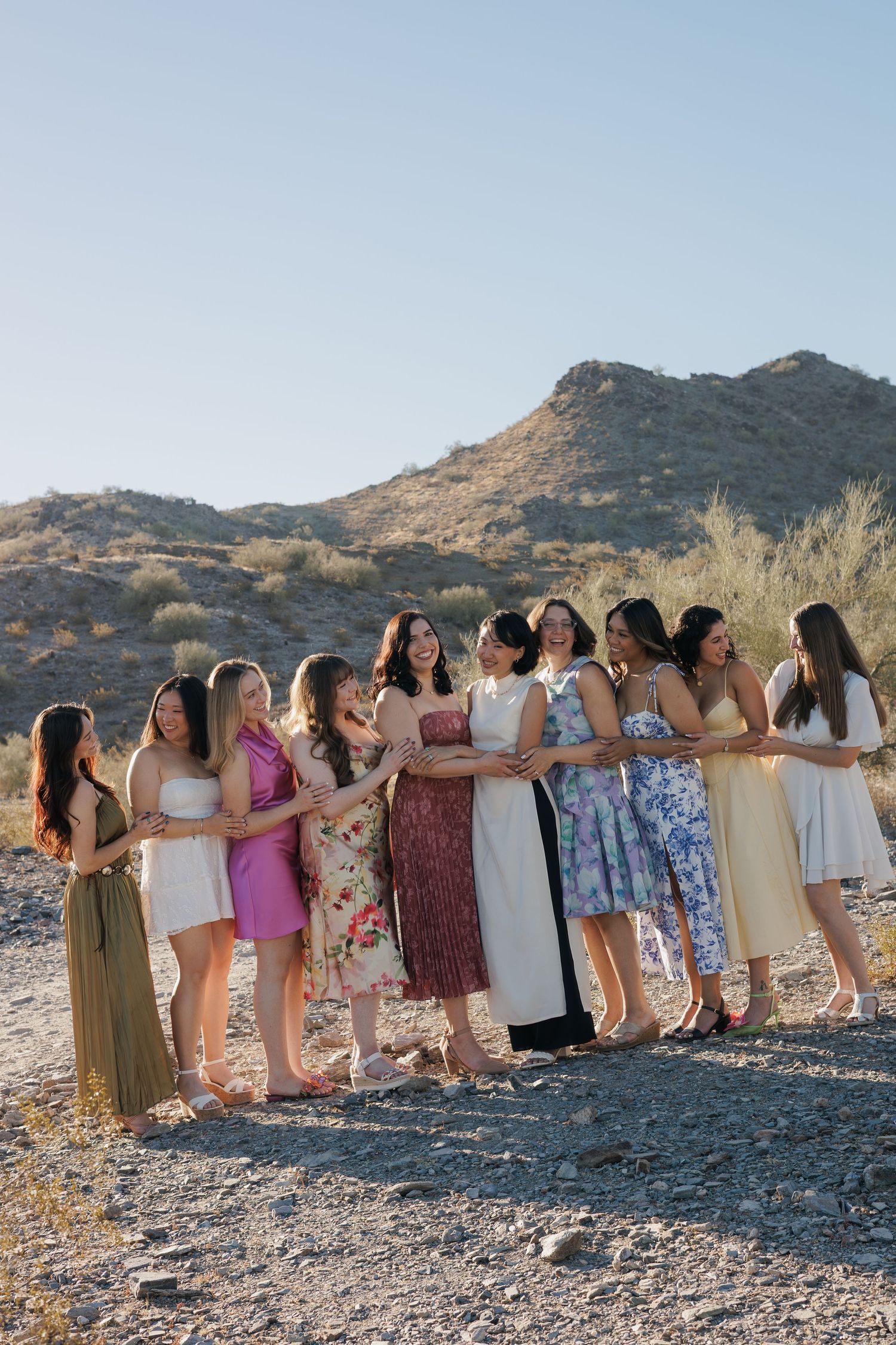 Group of women in colorful dresses posing together outdoors in a desert landscape with rocky hills in the background.