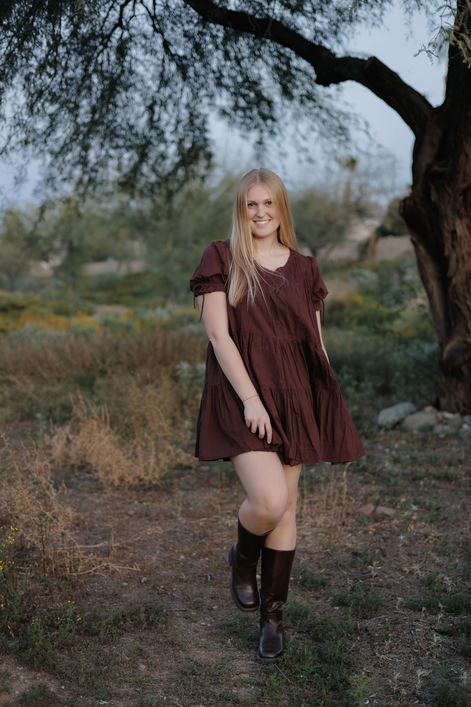 Person in flowing brown dress poses playfully in desert landscape under mesquite tree.