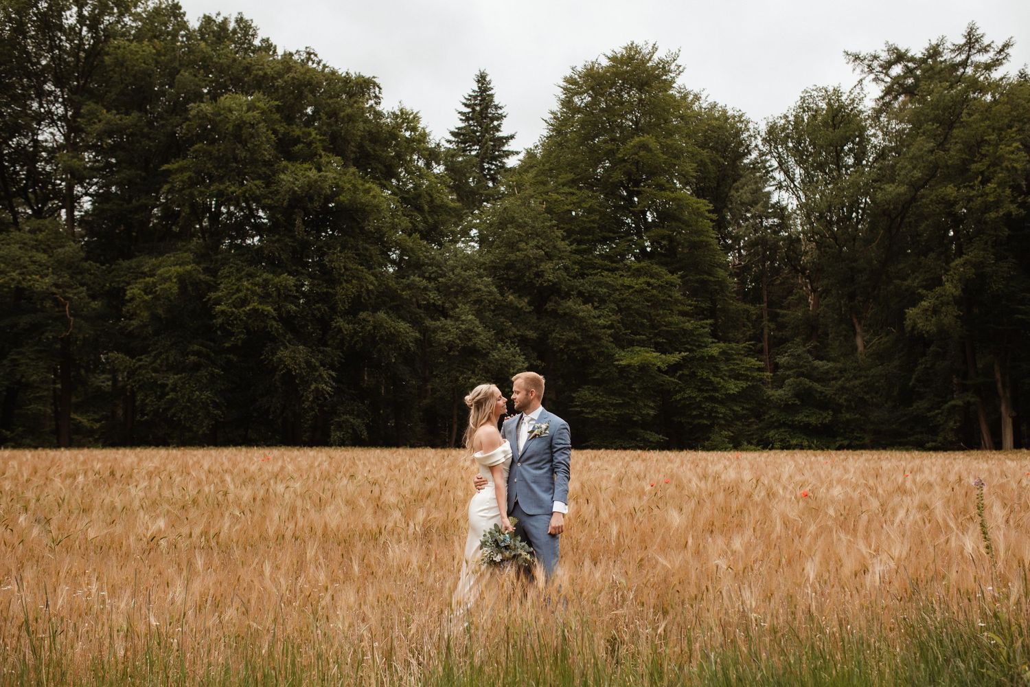 A couple stands together in a golden wheat field surrounded by trees.