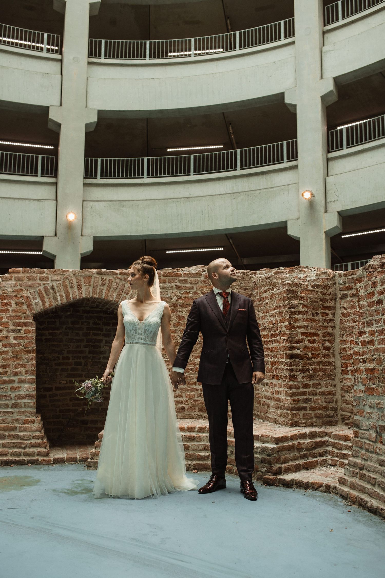 A couple poses in wedding attire within a curved architectural space with brick and concrete details.