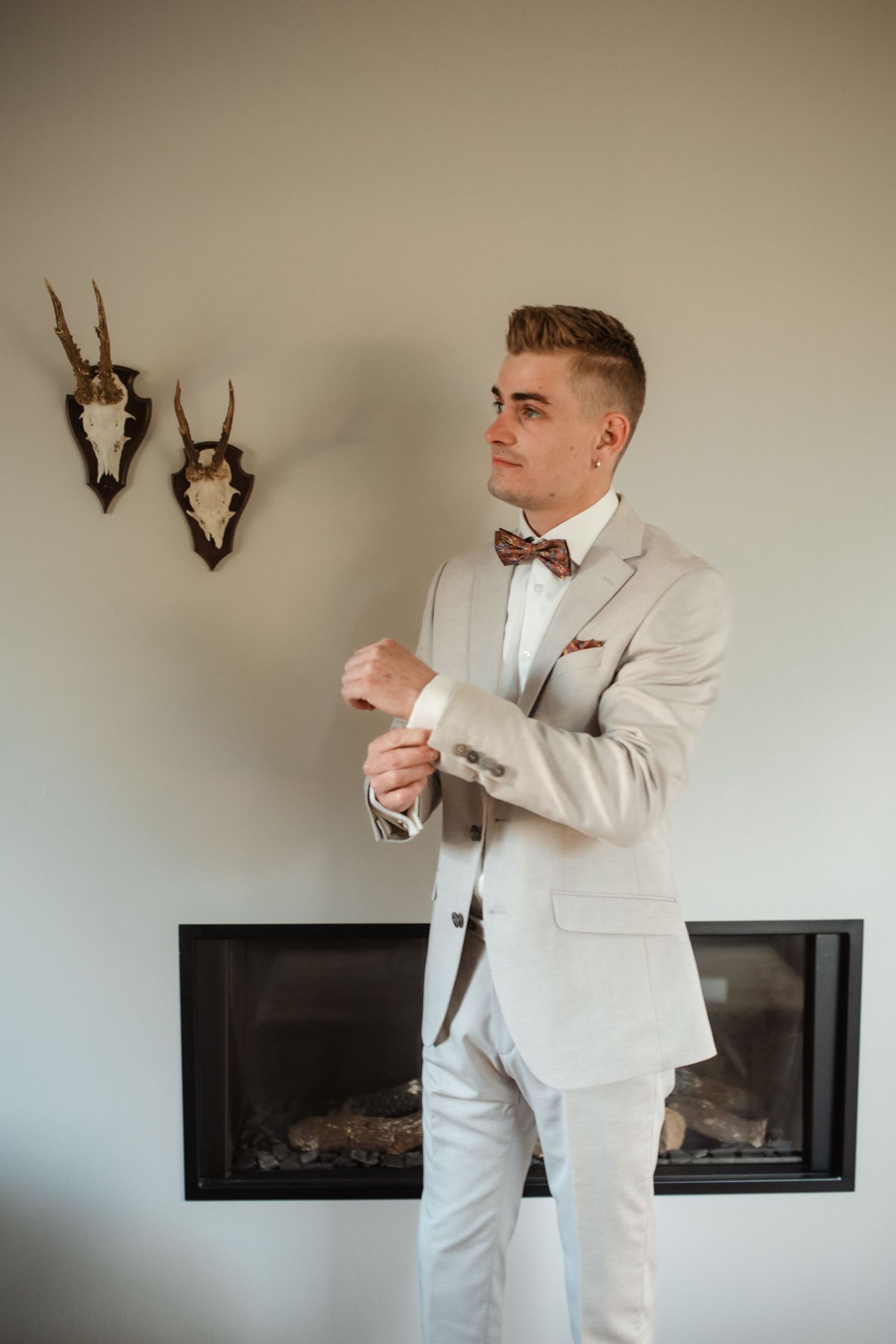Someone in a white suit and bow tie adjusts their attire in front of a modern fireplace.