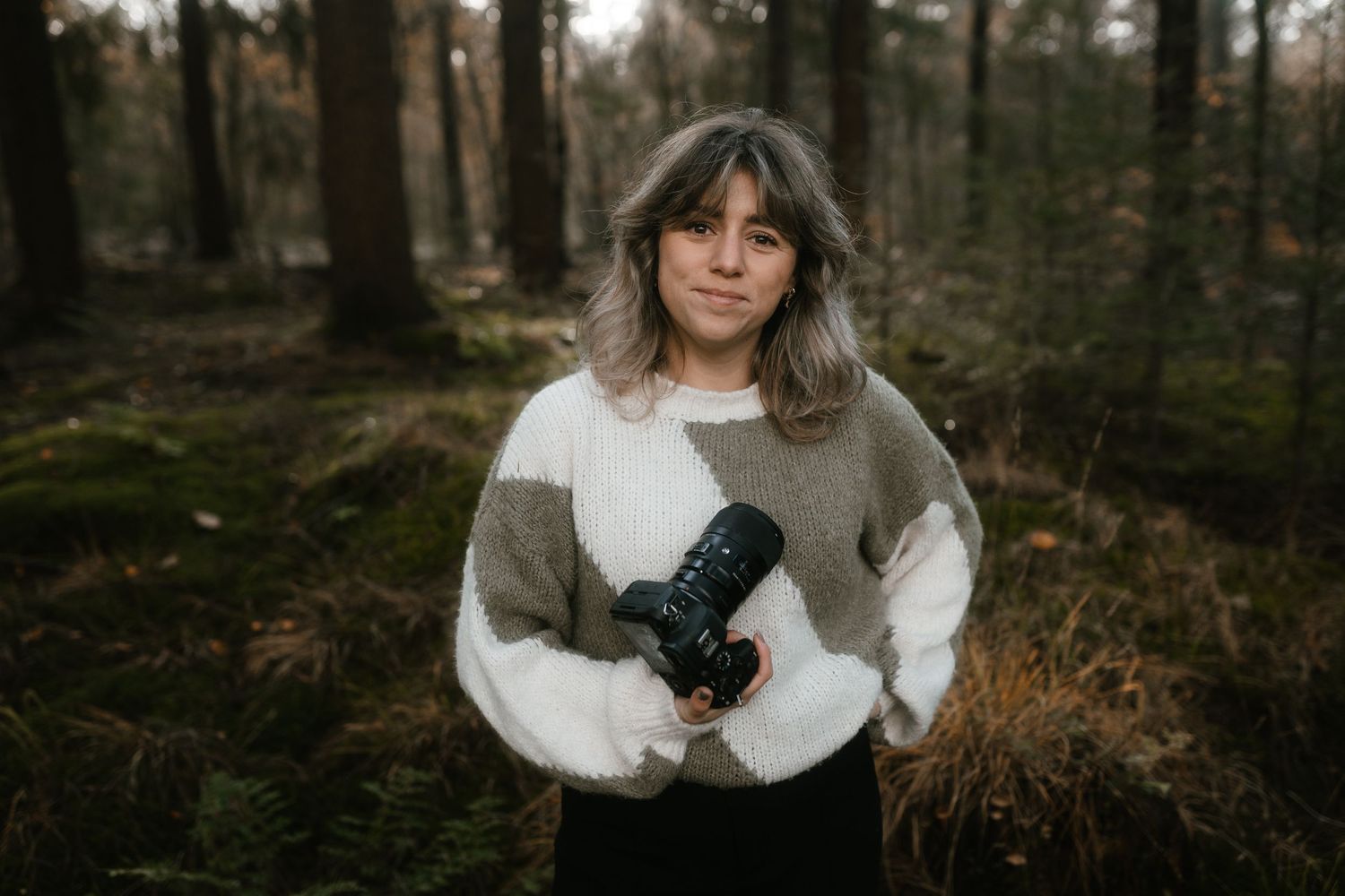 Portrait of photographer in forest wearing chunky knit sweater holding professional camera.