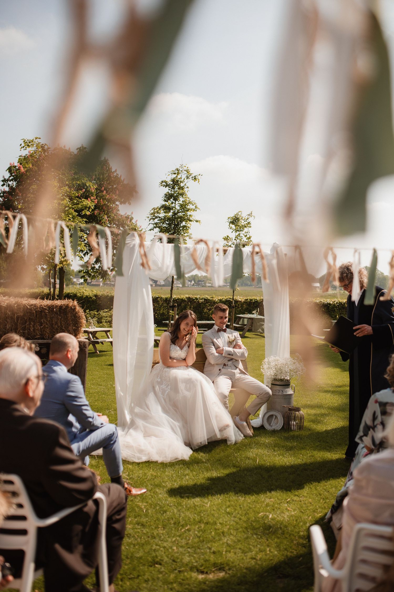 Guests seated at an outdoor wedding ceremony with white fabric draped posts and natural scenery.