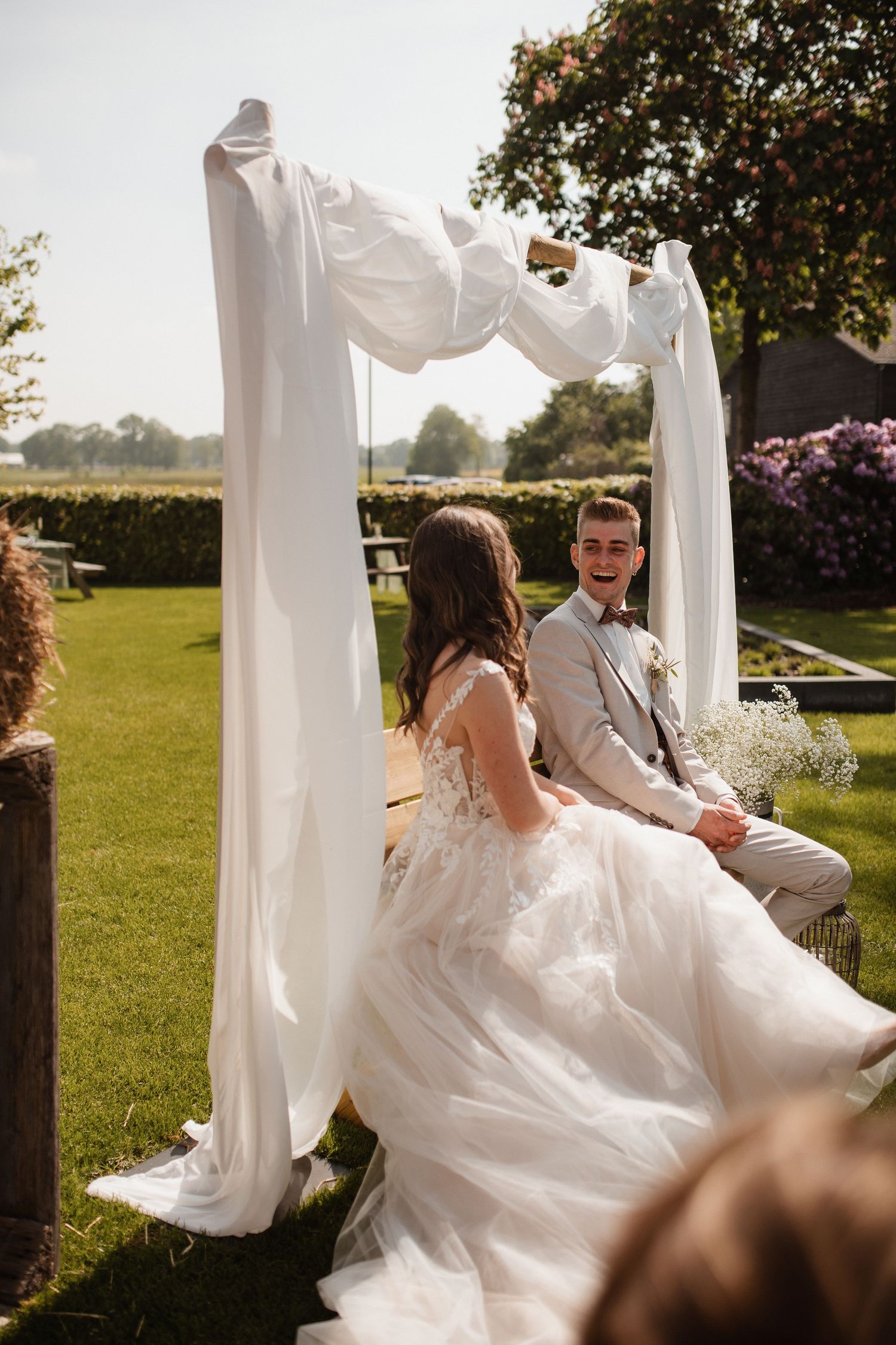 A couple sits on wooden benches under flowing white fabric drapes in an outdoor garden setting.