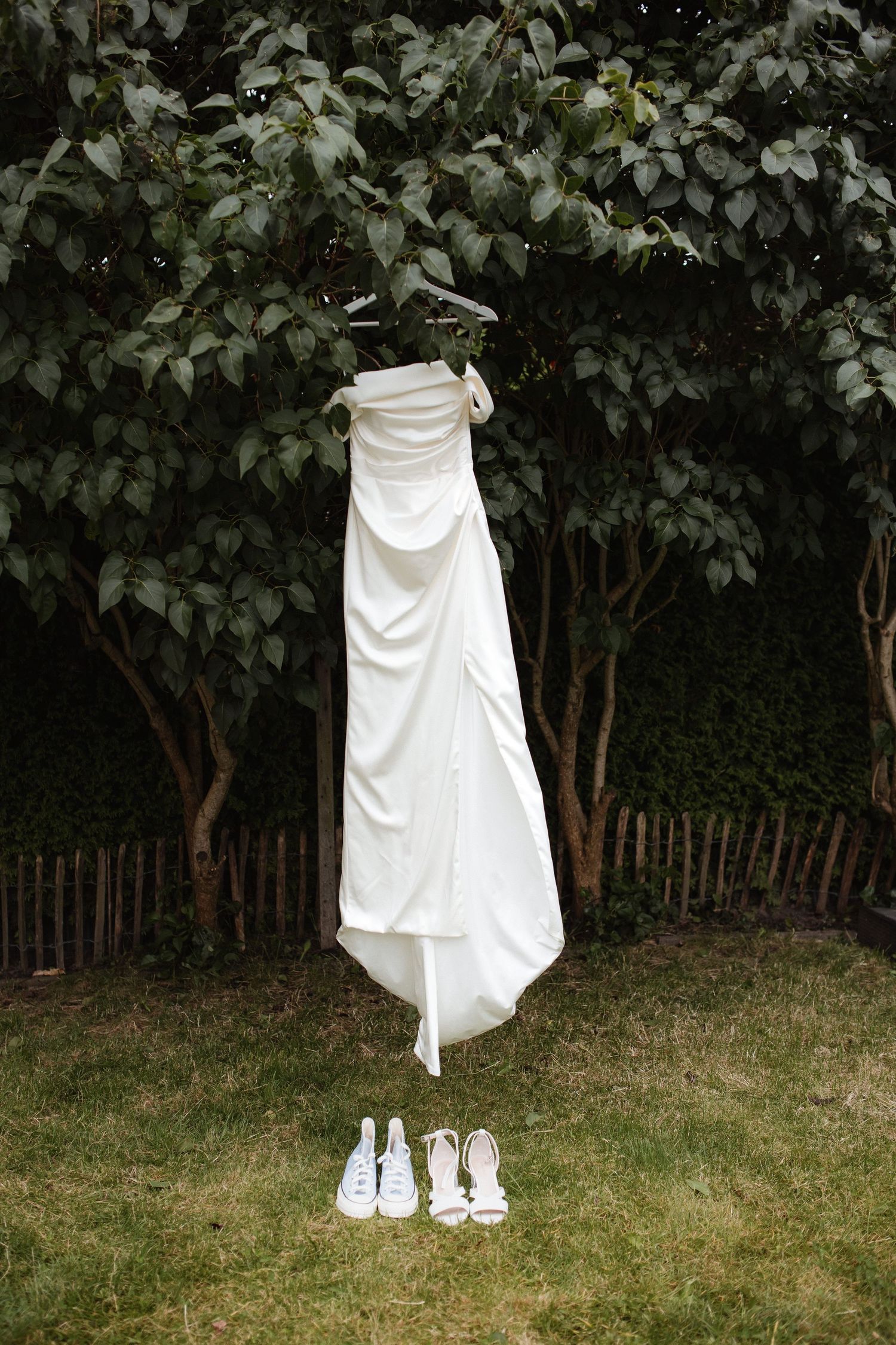 White wedding dress hanging on a tree with two pairs of white shoes beneath it.