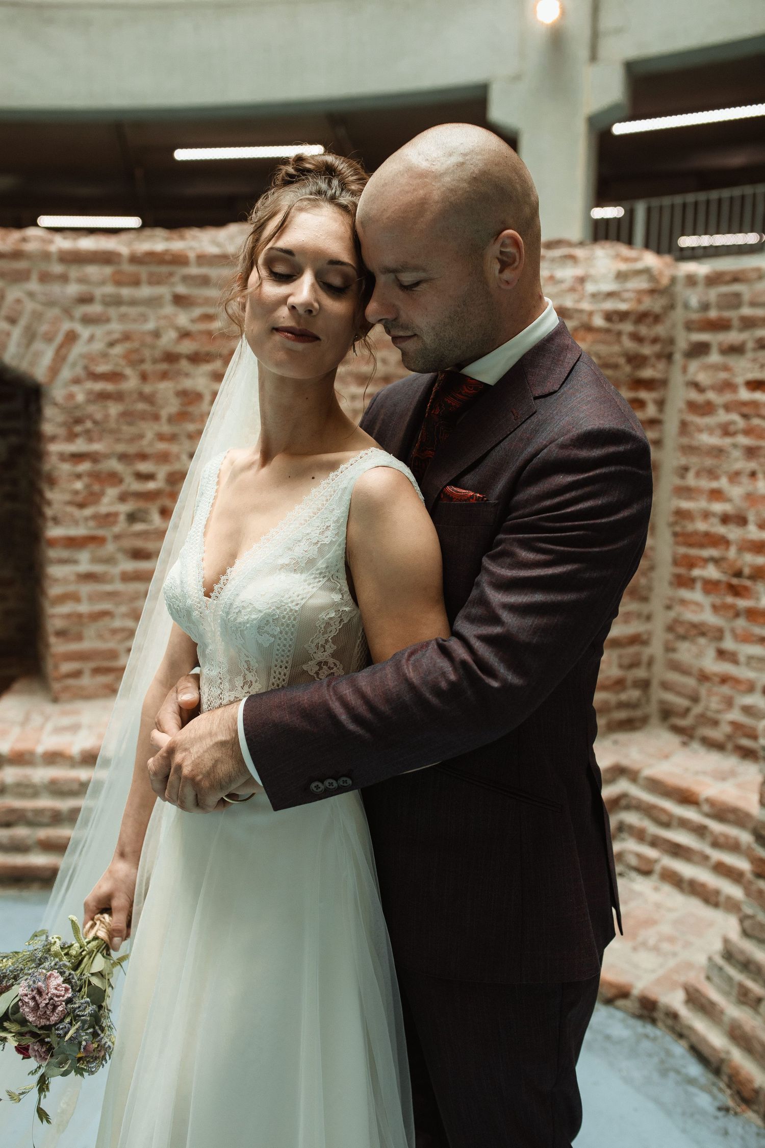 A couple in wedding attire shares a tender moment against an exposed brick wall background.