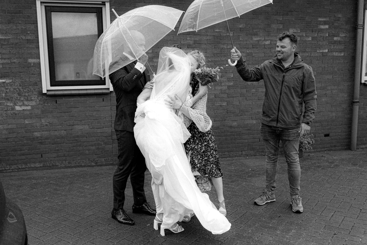 A wedding party huddles under clear umbrellas on a rainy day by a brick building.