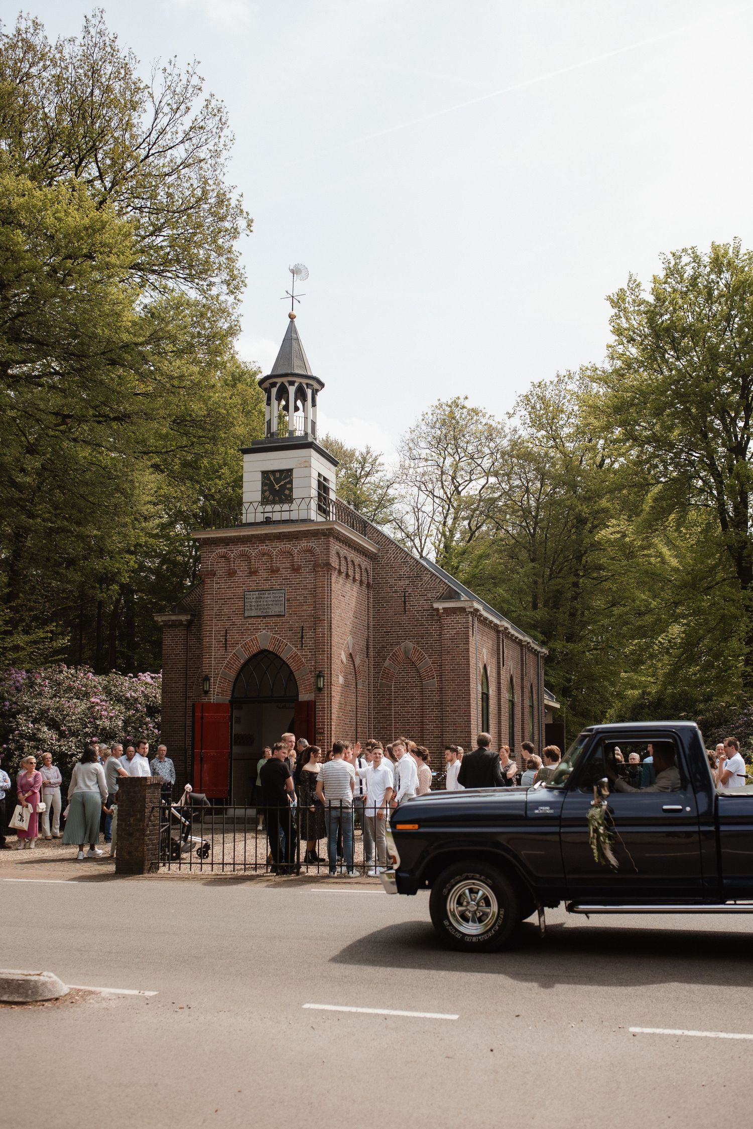 A classic brick church with a steeple and red doors stands among trees as vintage cars arrive for a wedding ceremony.