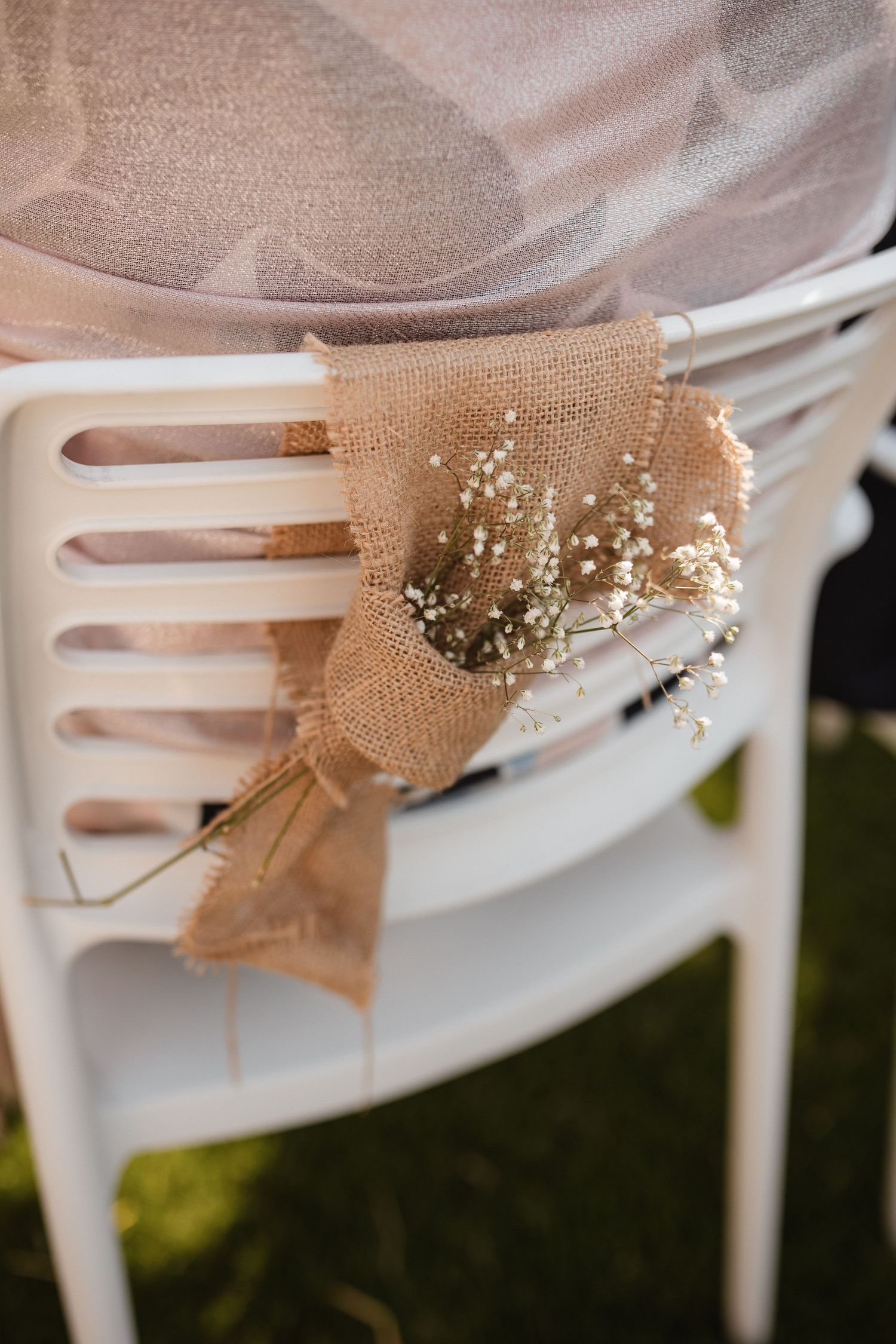 Rustic burlap bows with delicate baby's breath flowers adorn white chairs at an outdoor event.