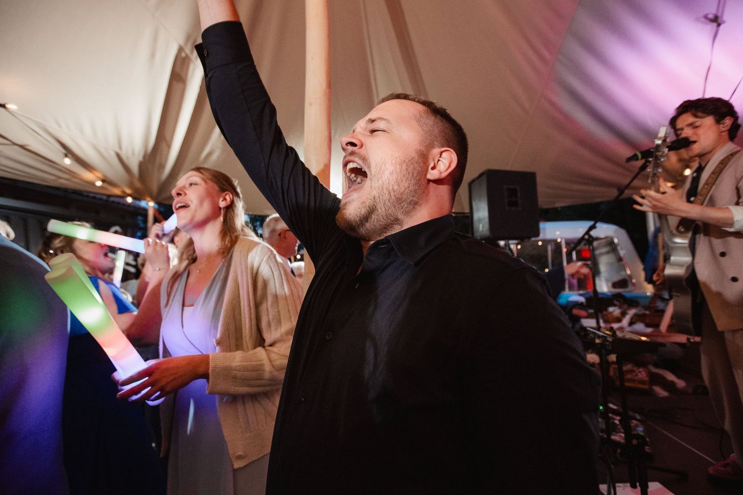 Party guests sing and dance enthusiastically under colorful lighting at an indoor celebration.