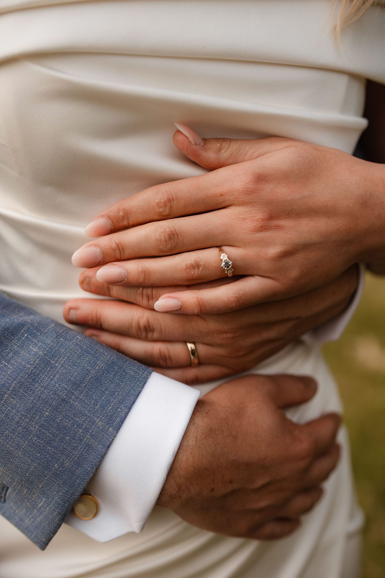Close-up details of hands wearing wedding rings against white fabric and blue suit.