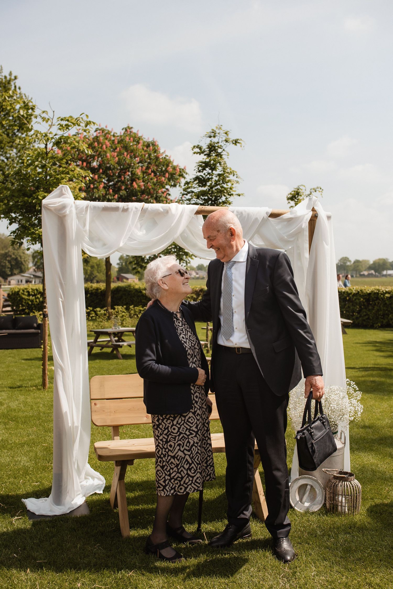 Elderly guests chat near a white fabric wedding arch in a sunny garden setting.