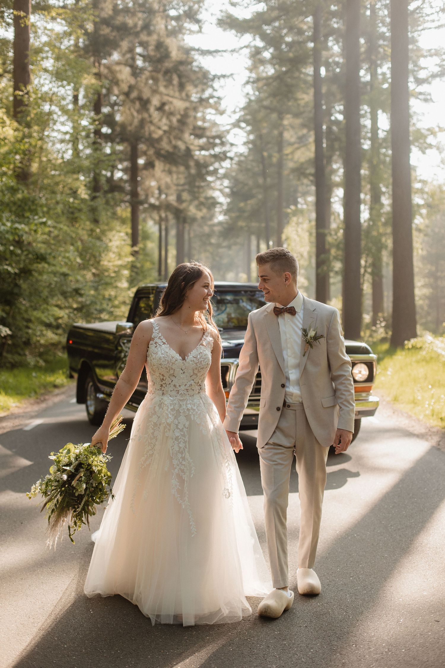 A wedding couple walks along a sunlit forest path surrounded by tall evergreen trees.