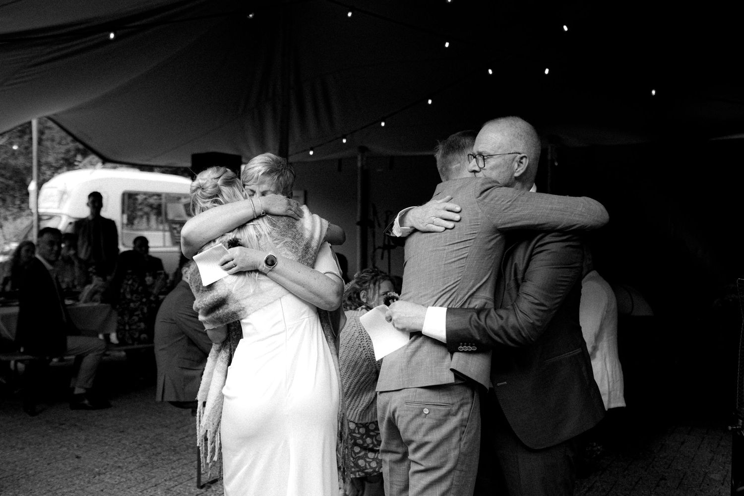 Black and white photo of emotional embraces during celebration under tent with twinkling lights.
