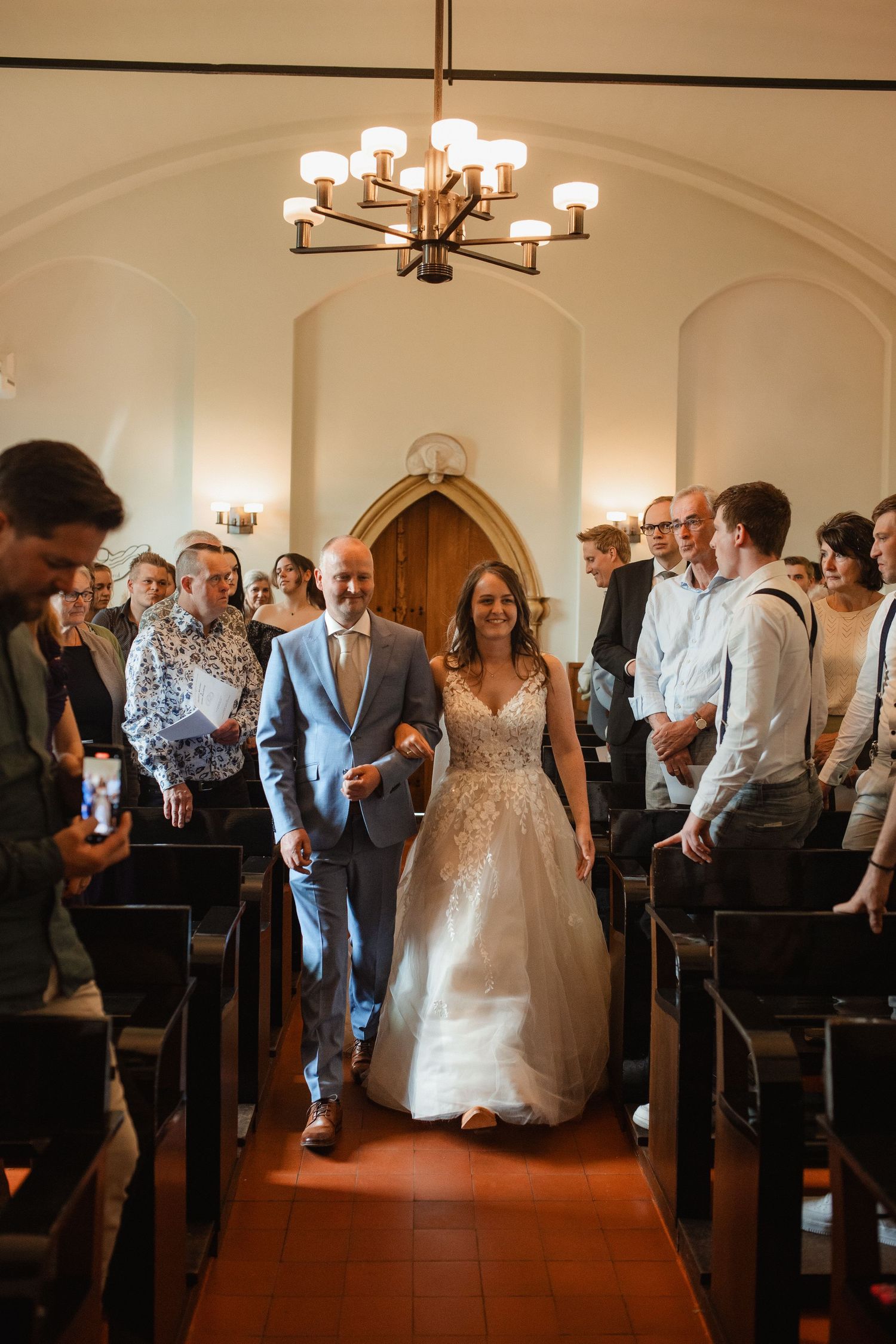 Wedding guests gather in church pews under elegant chandeliers during a ceremony.