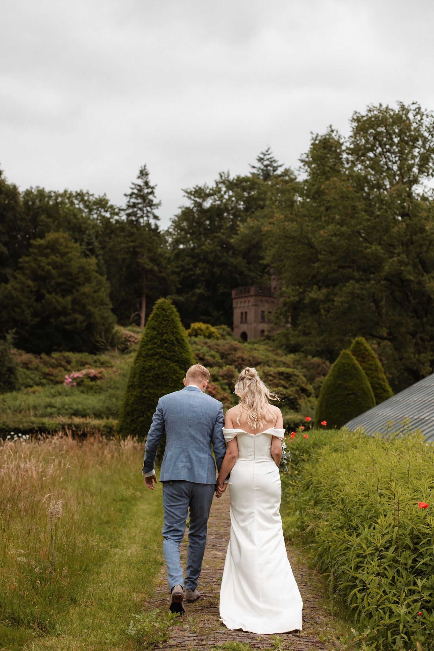 A couple walks through a garden path towards a stone building on their wedding day.