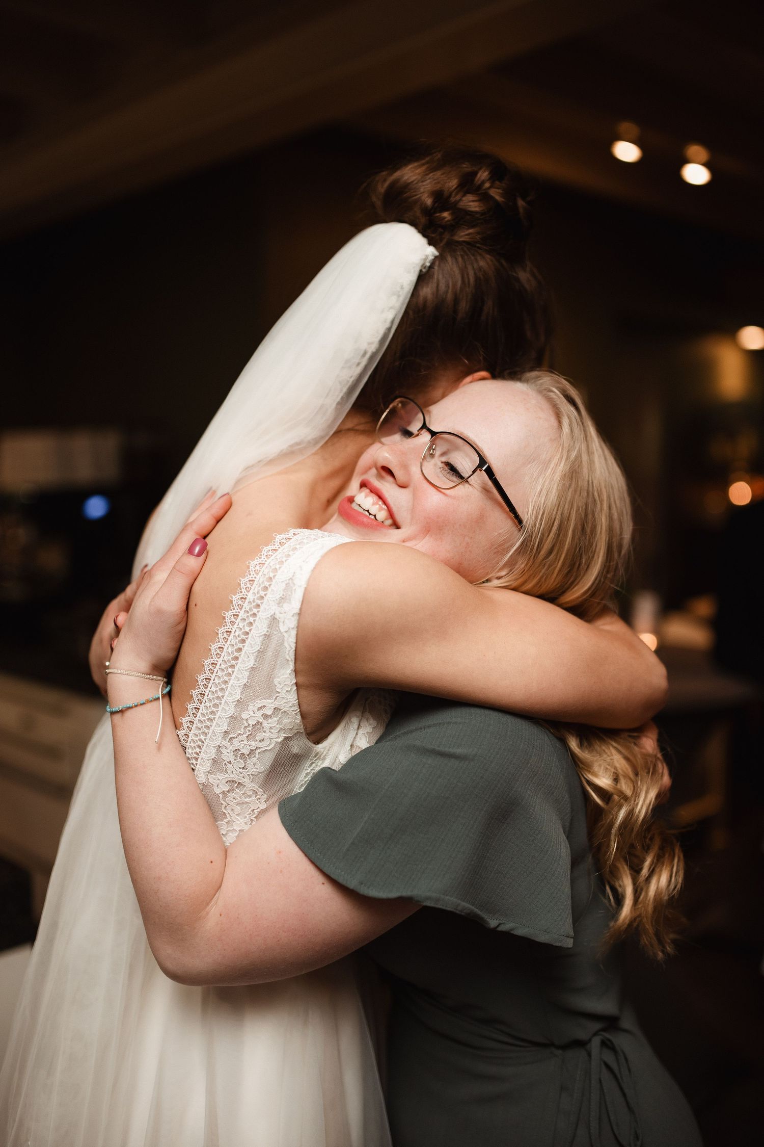 Bride in white lace dress and veil shares emotional embrace during wedding reception.