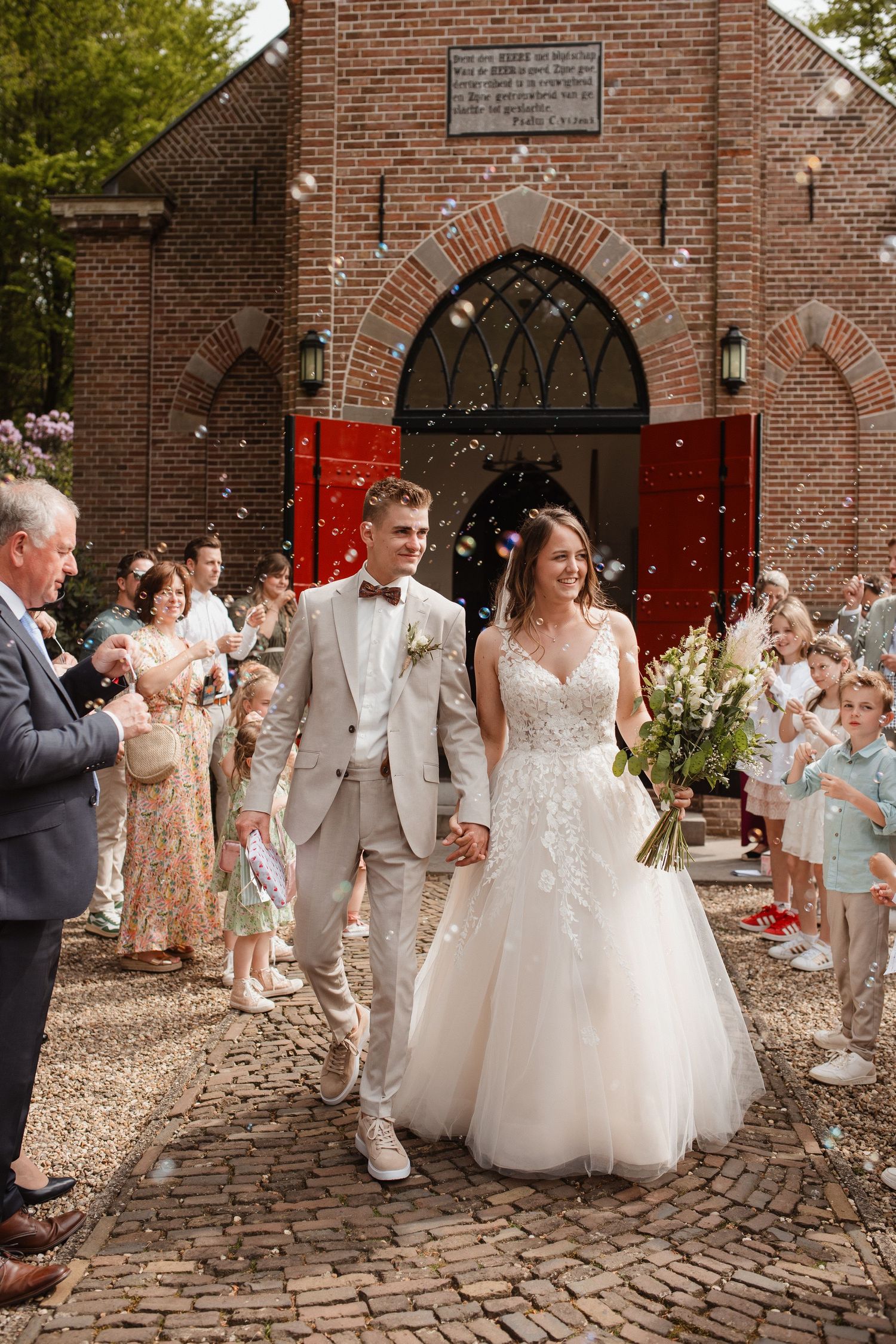 Wedding party exits brick church as guests throw confetti on cobblestone path.