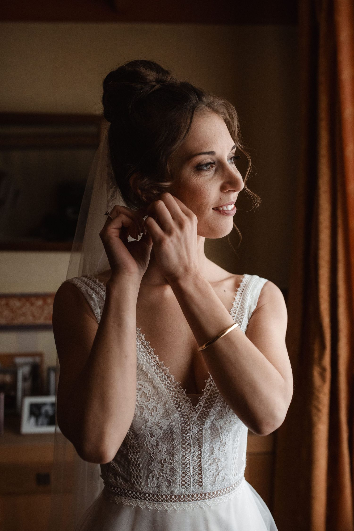 A bride adjusts her jewelry while wearing an elegant lace wedding dress.