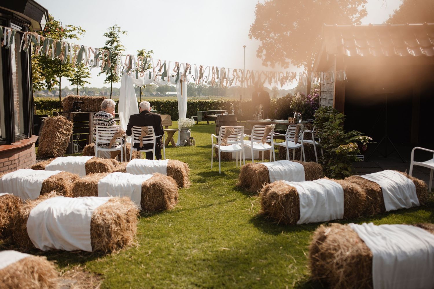 Rustic outdoor wedding reception setup with hay bales and white linens in a garden setting.