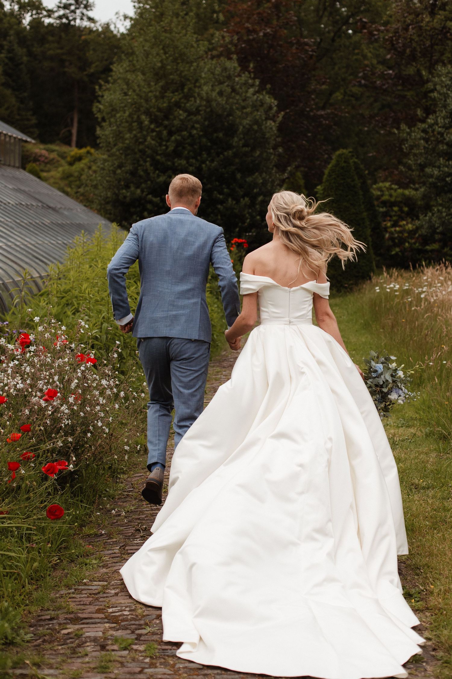 A couple walking down a garden path with red roses, the bride wearing a flowing white off-shoulder gown.