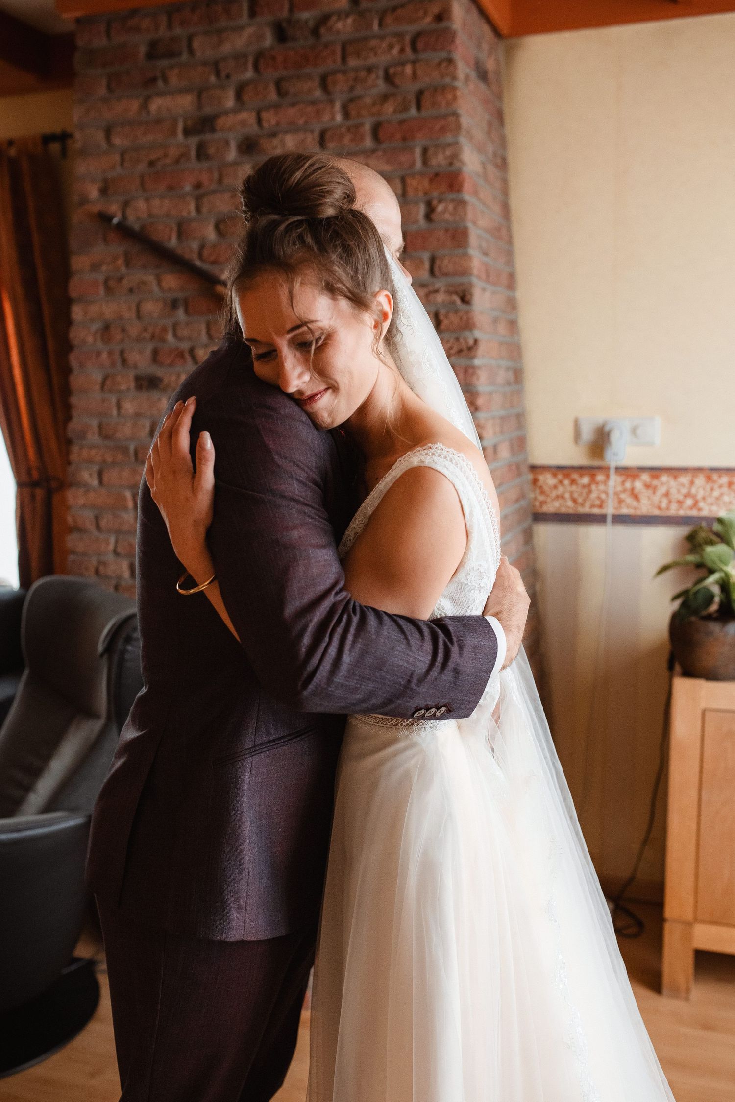 Two people embrace emotionally in wedding attire inside a room with exposed brick walls.