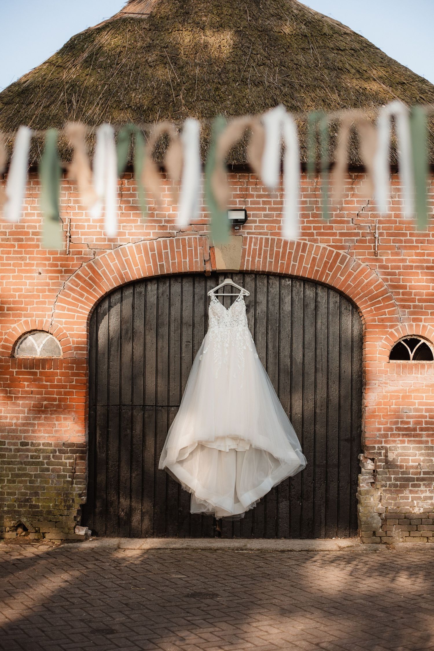 A white wedding dress hangs against rustic brick walls with black wooden doors.