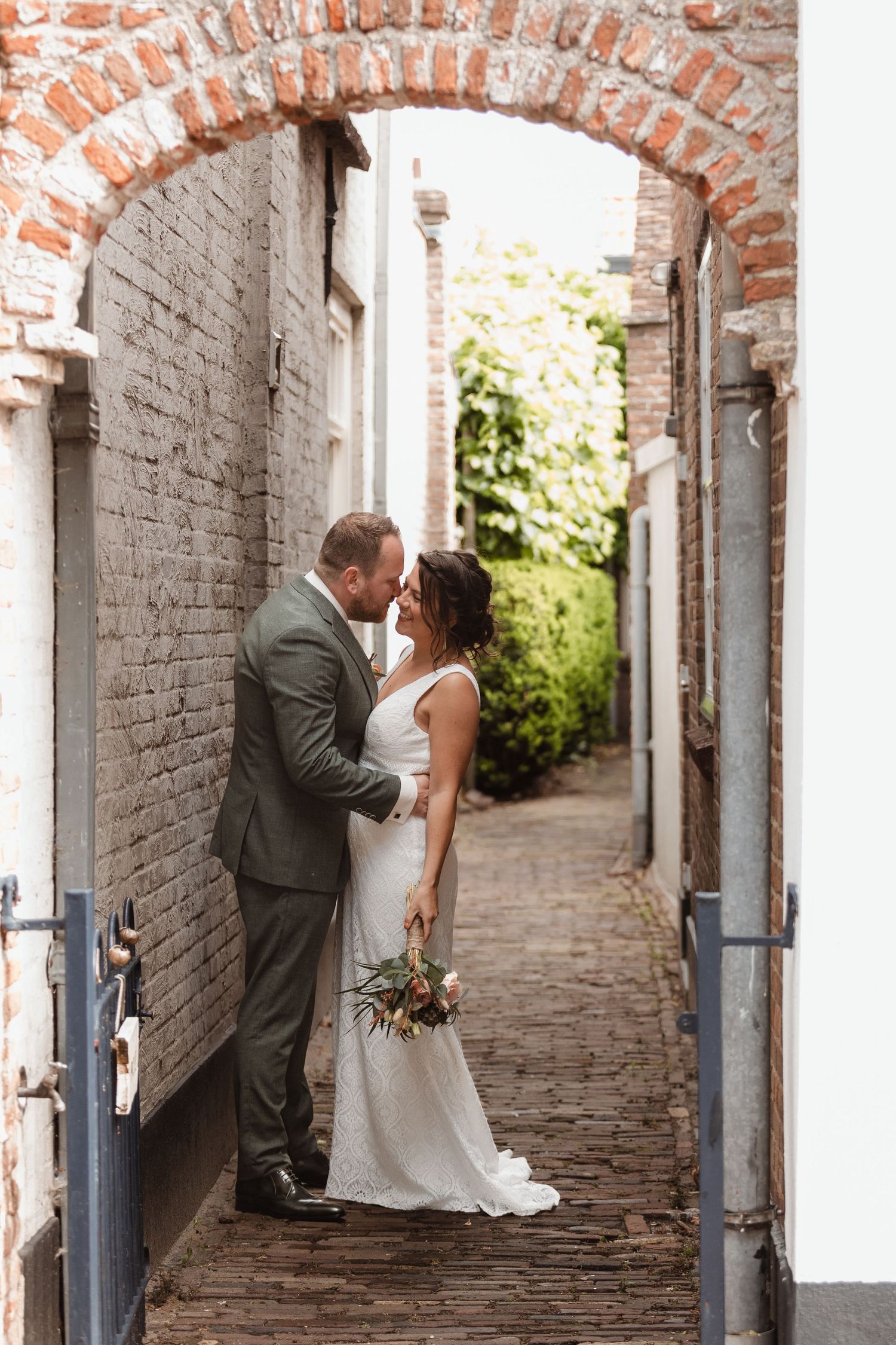 Wedding couple poses under brick archway in historic alleyway.