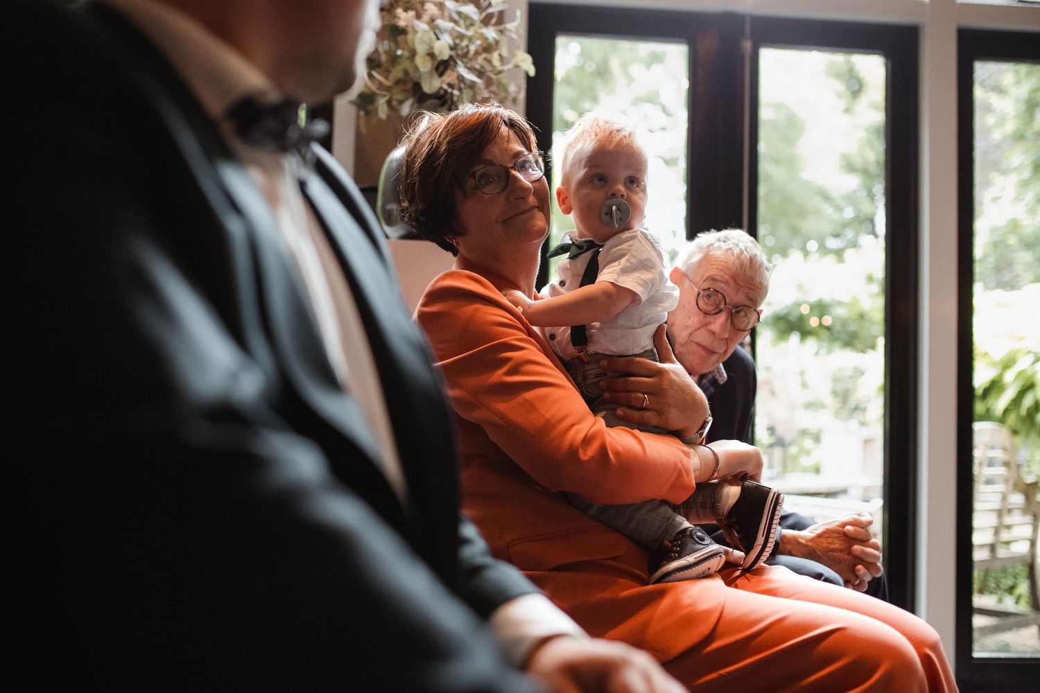 Wedding guests in orange attire seated by large windows during ceremony.