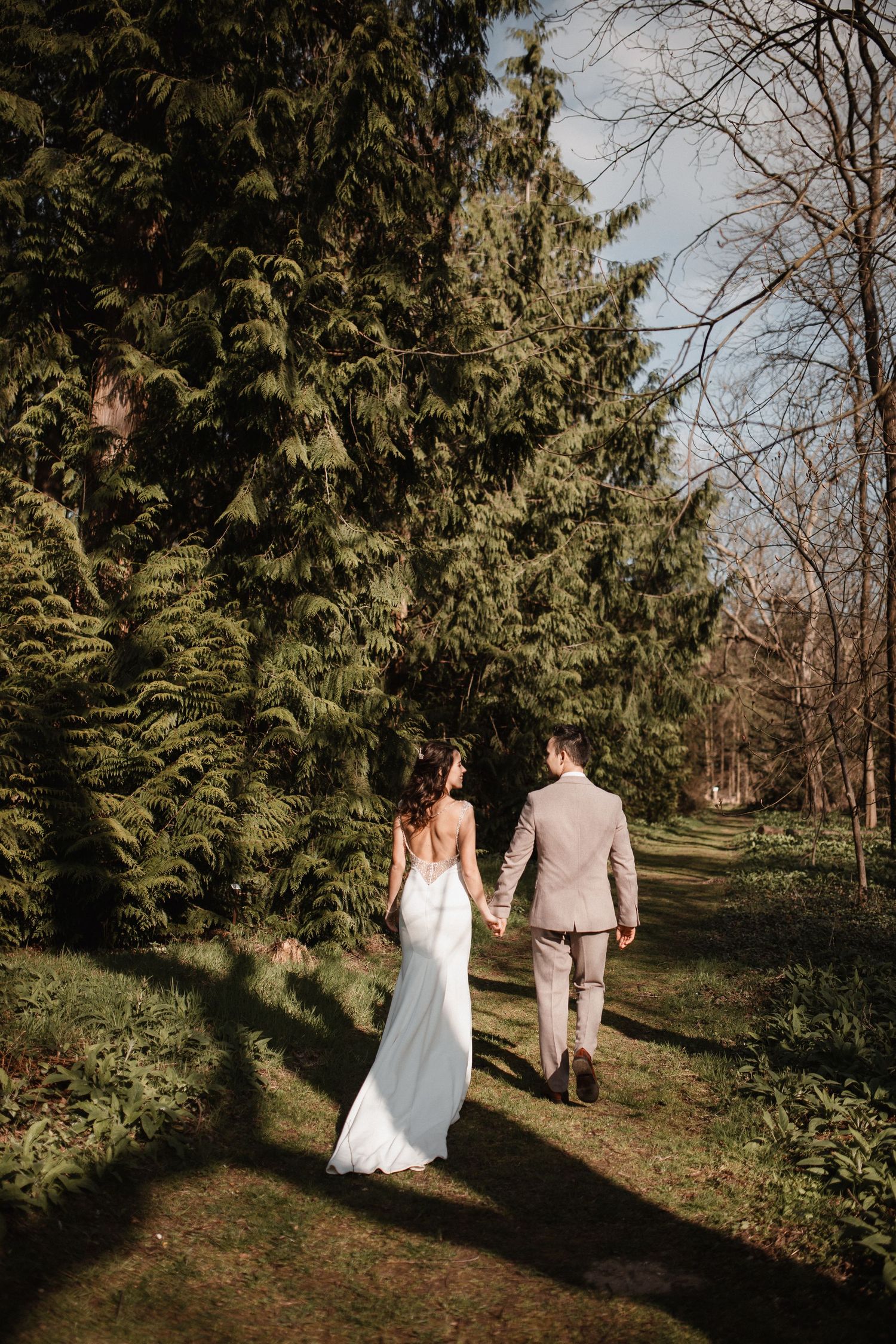 Wedding couple walking through a scenic wooded path with evergreen trees on a sunny day.