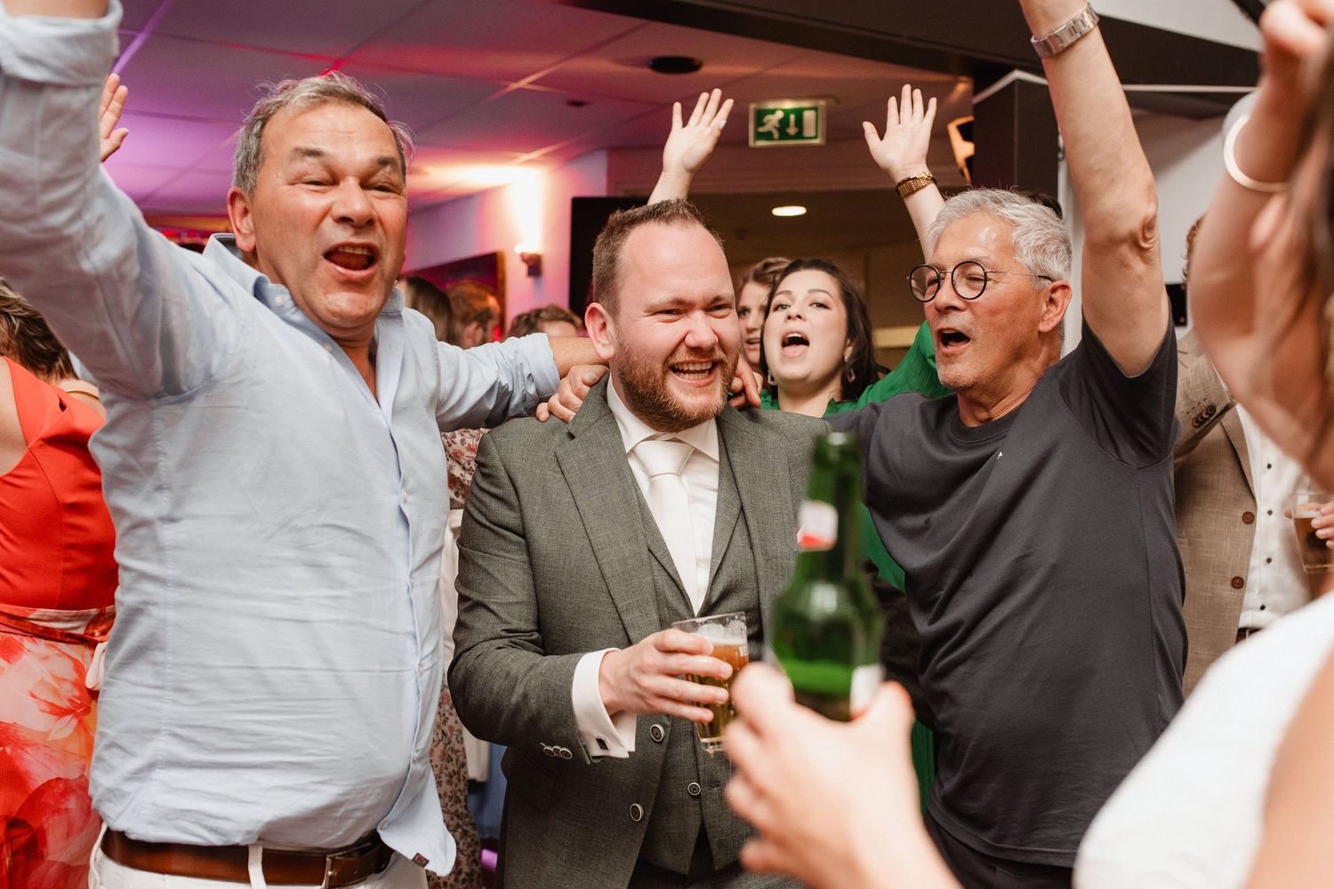 Lively party crowd celebrating with drinks and raised hands in a dimly lit venue.