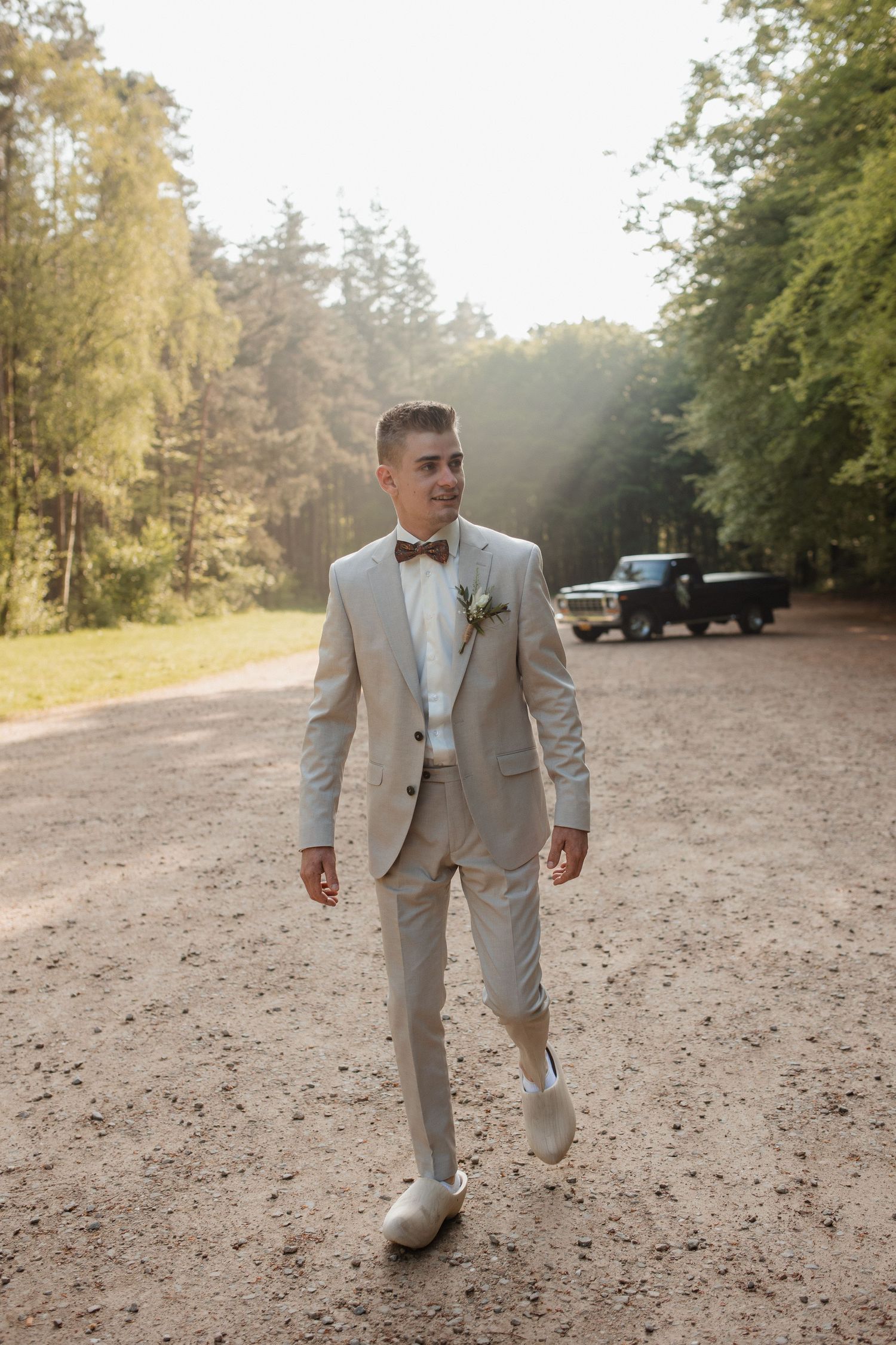 A groom in a light gray suit walks across a dirt path surrounded by trees on a sunny day.