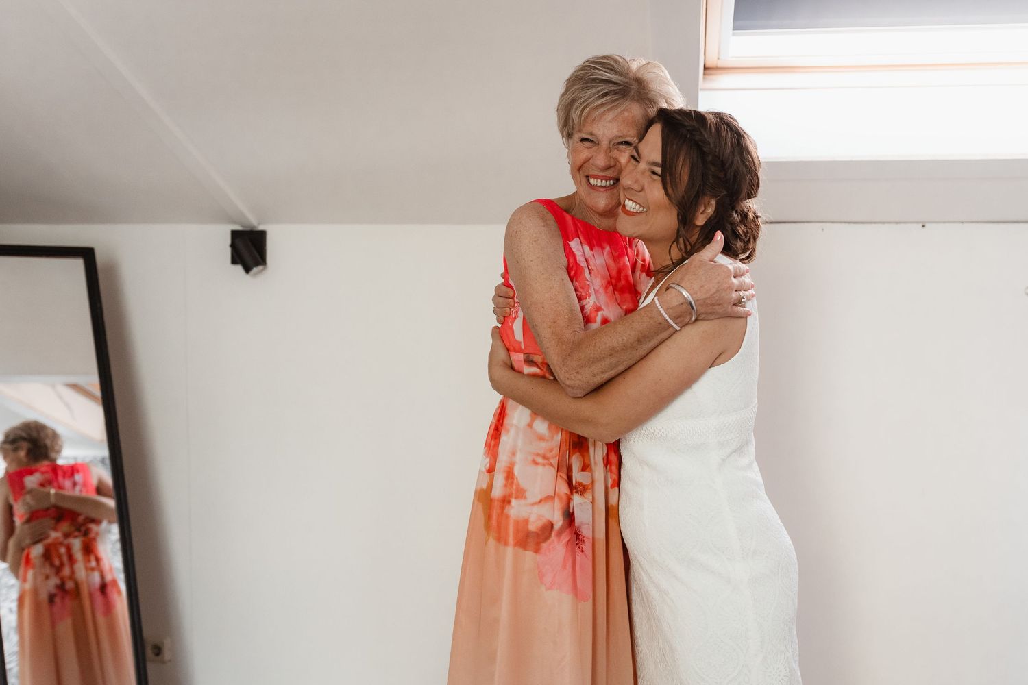 Bride in a coral dress puts on shoes in a room with floral wallpaper.