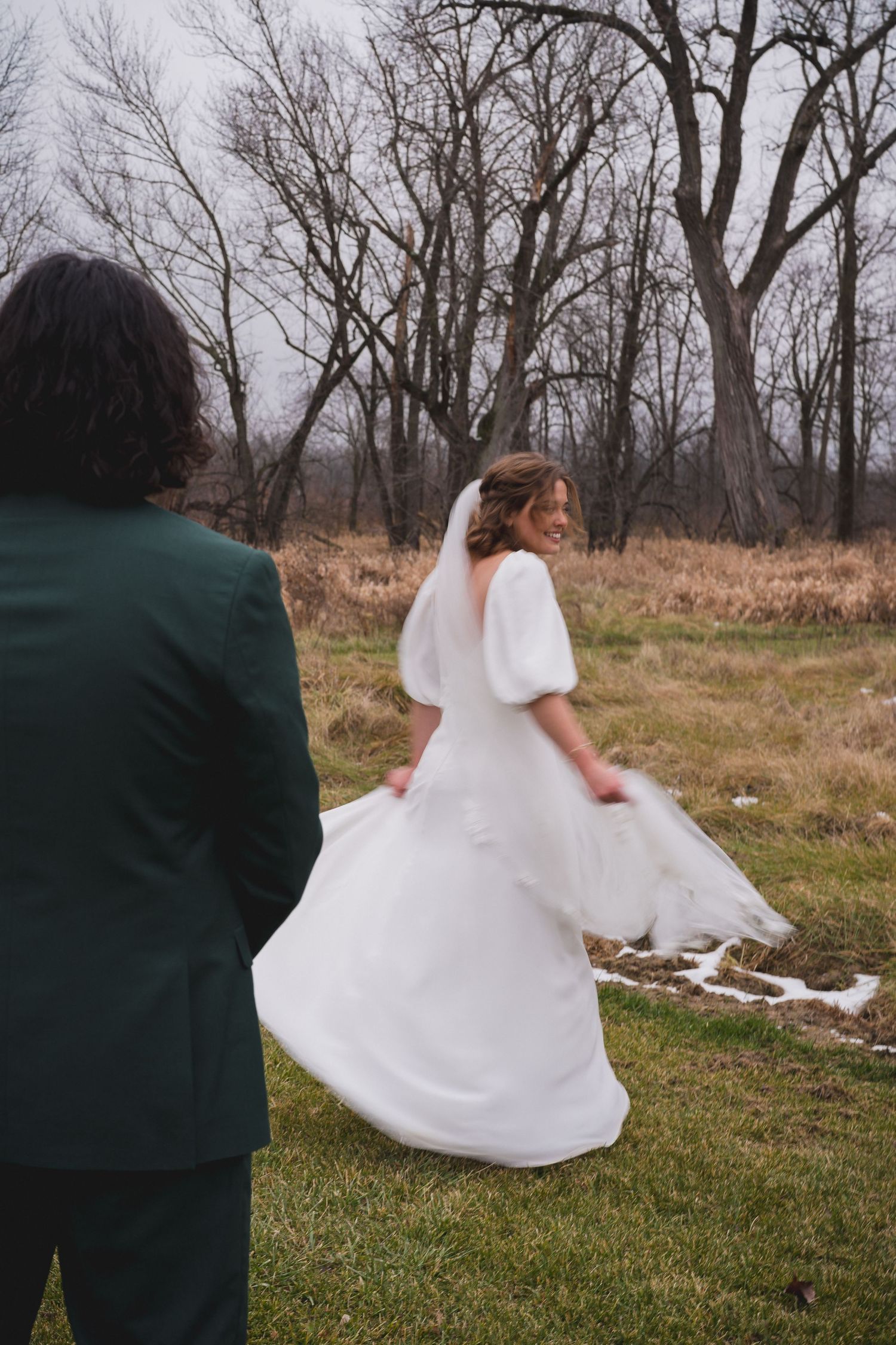 A wedding couple poses in a winter field with dried grass and bare trees.