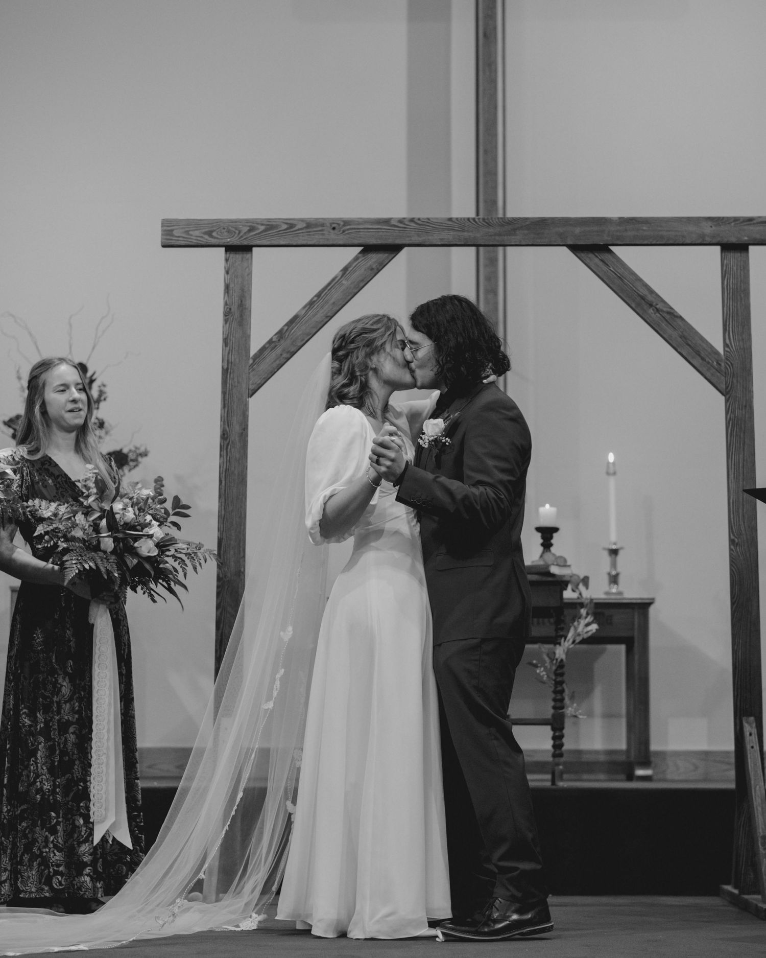 Black and white photo captures first kiss during wedding ceremony beneath wooden cross.