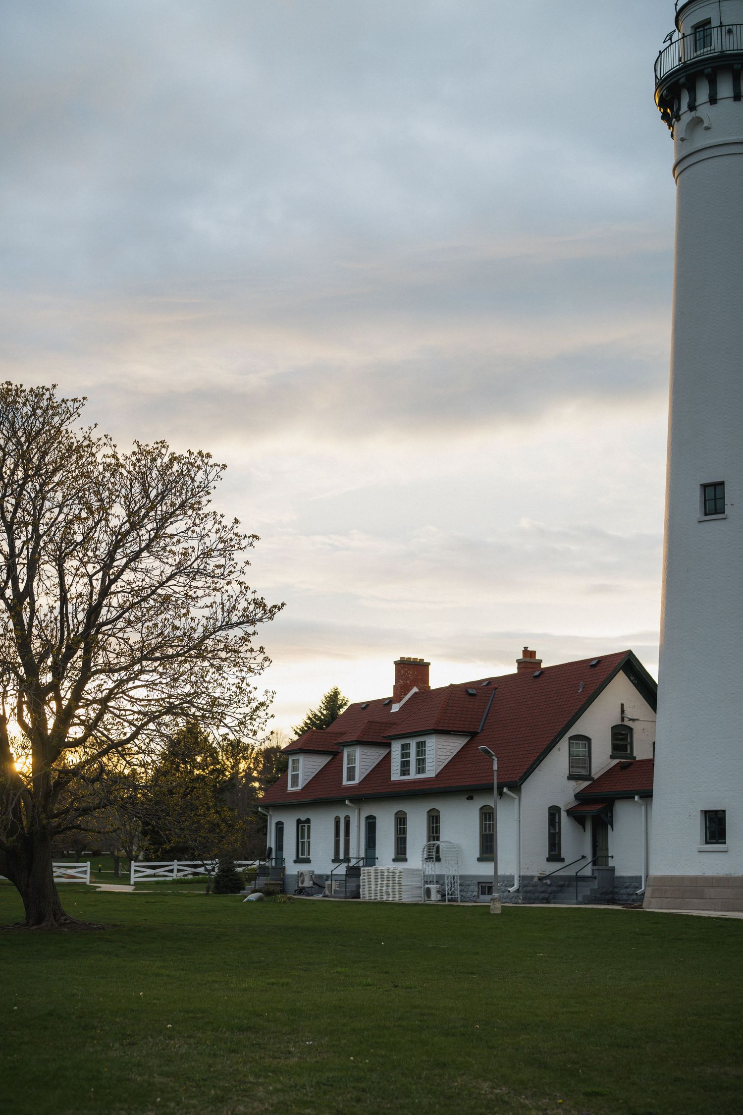 White lighthouse tower beside a red-roofed keeper's house at sunset, surrounded by green lawn and trees.