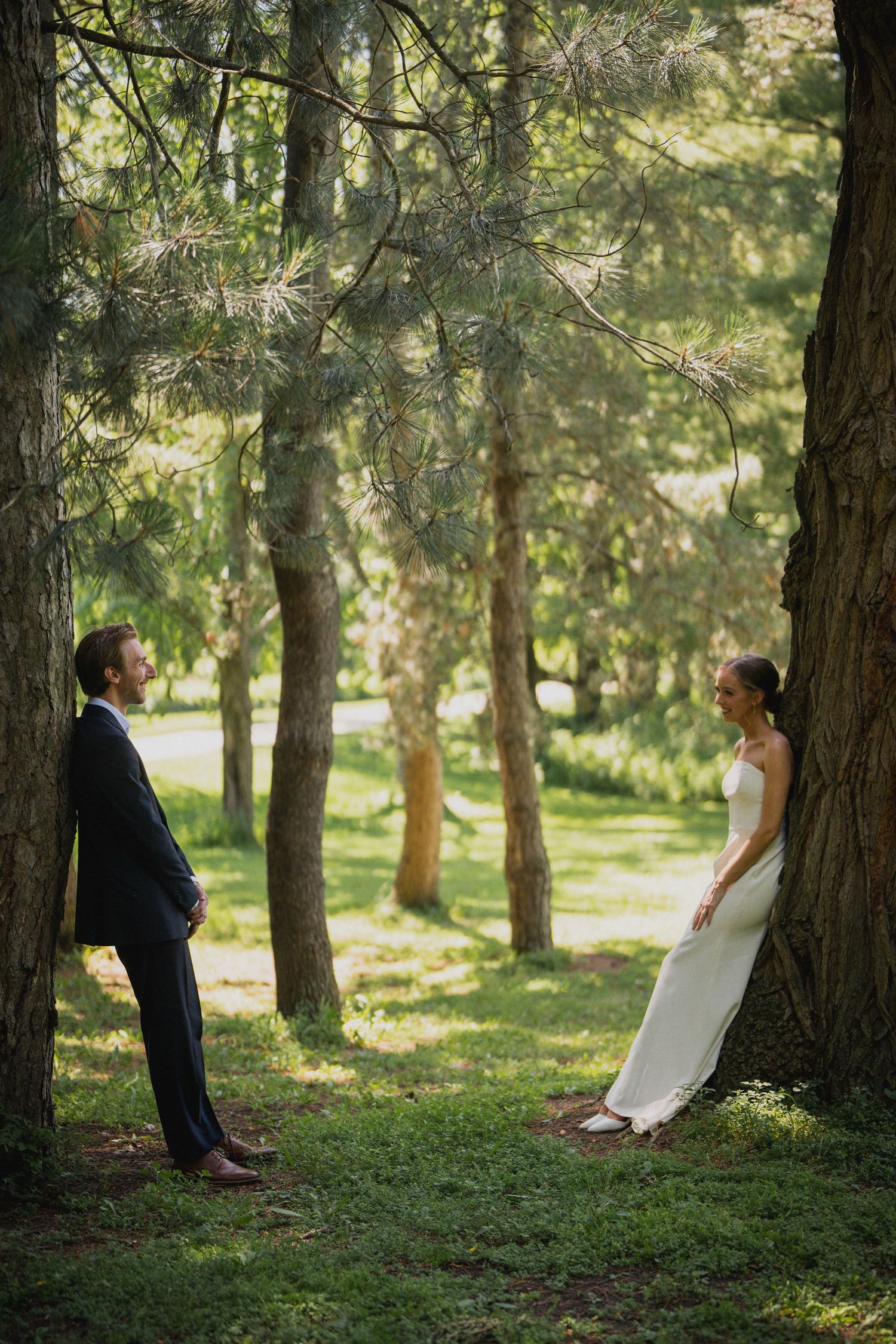 Bride and groom in wedding attire stand apart leaning against pine trees in a sunlit forest.
