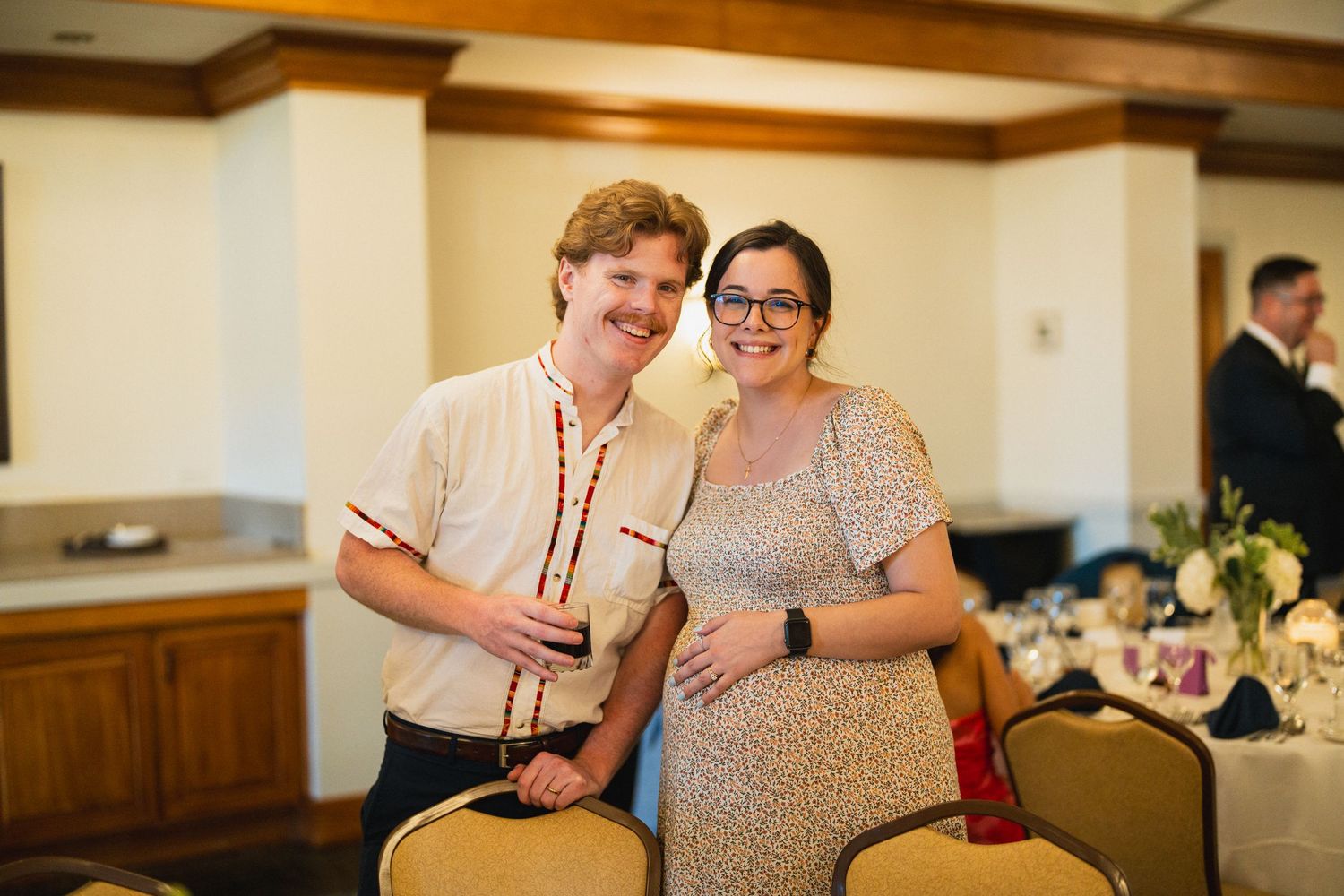 Two wedding guests smiling together inside a reception venue, woman in floral dress and man in retro shirt.