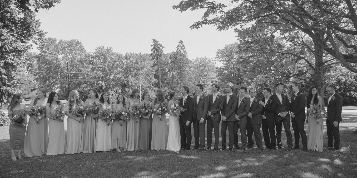 Large wedding party posing together outdoors under trees in black and white photo.