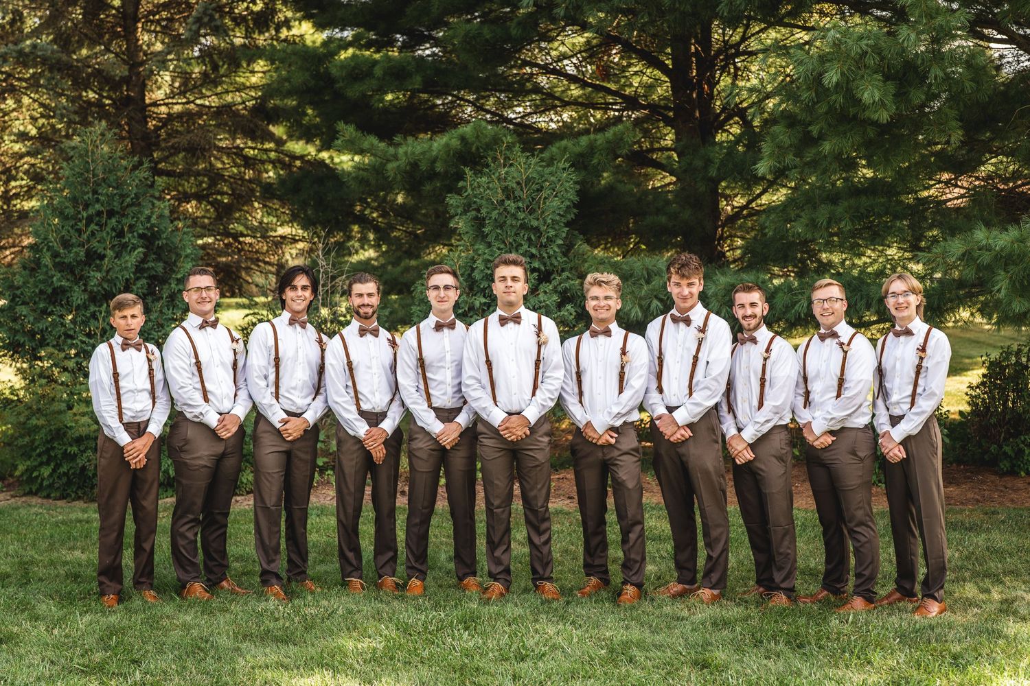 Groomsmen in matching brown pants and suspenders pose in line outdoors.