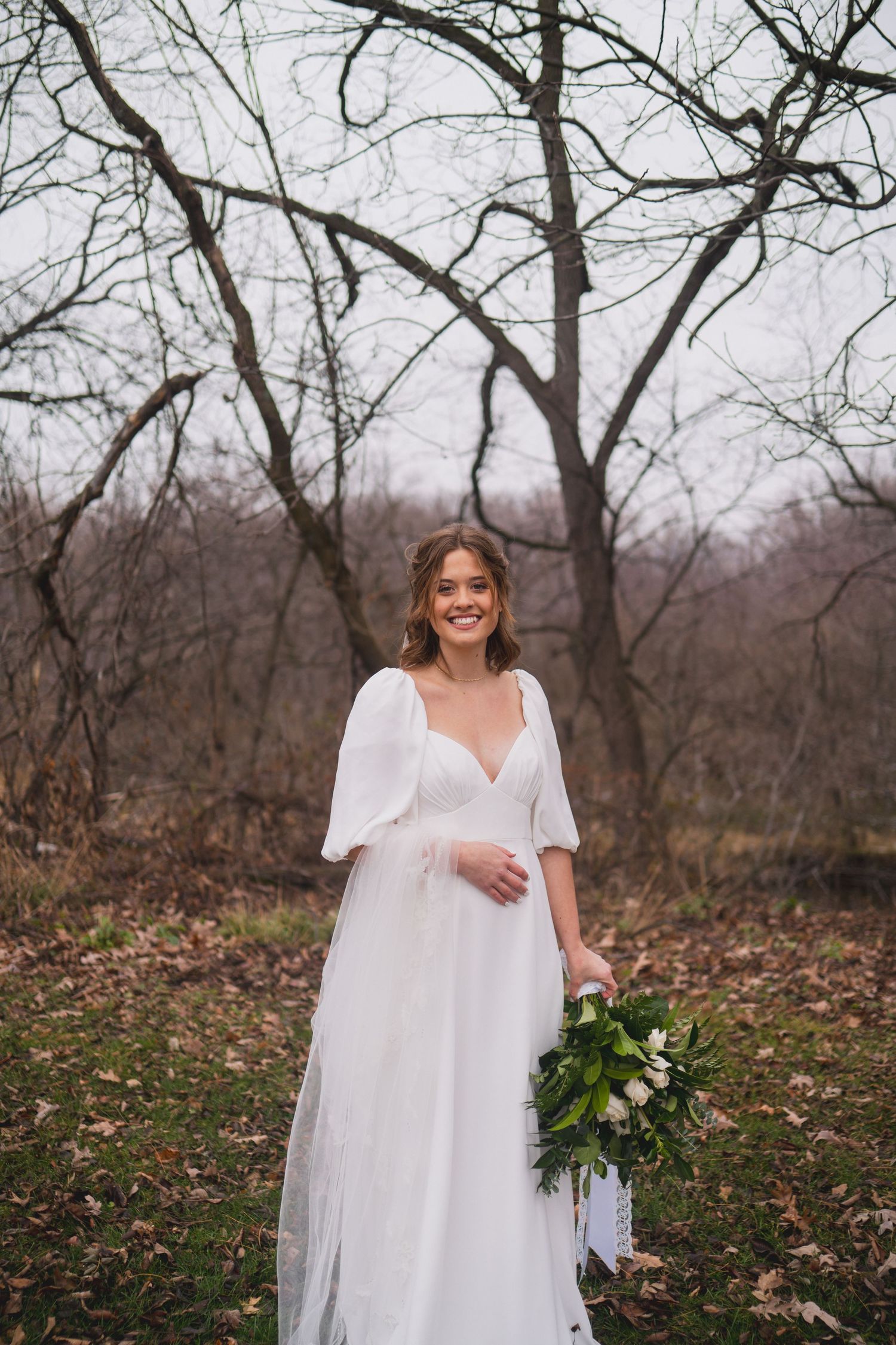 Full-length portrait of a bride in white dress holding bouquet against bare winter trees.