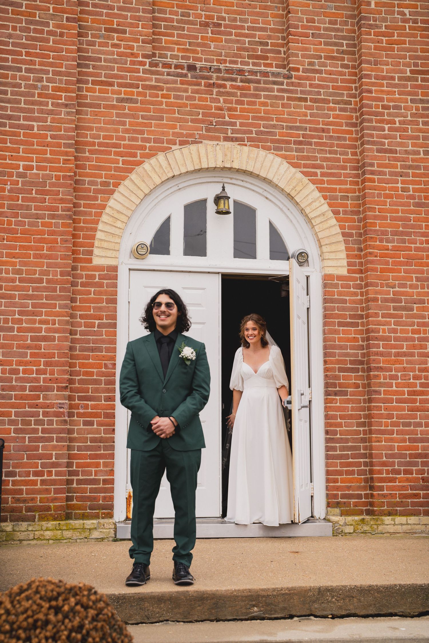 A couple in wedding attire stands in front of a brick church entrance with white arched doorways.