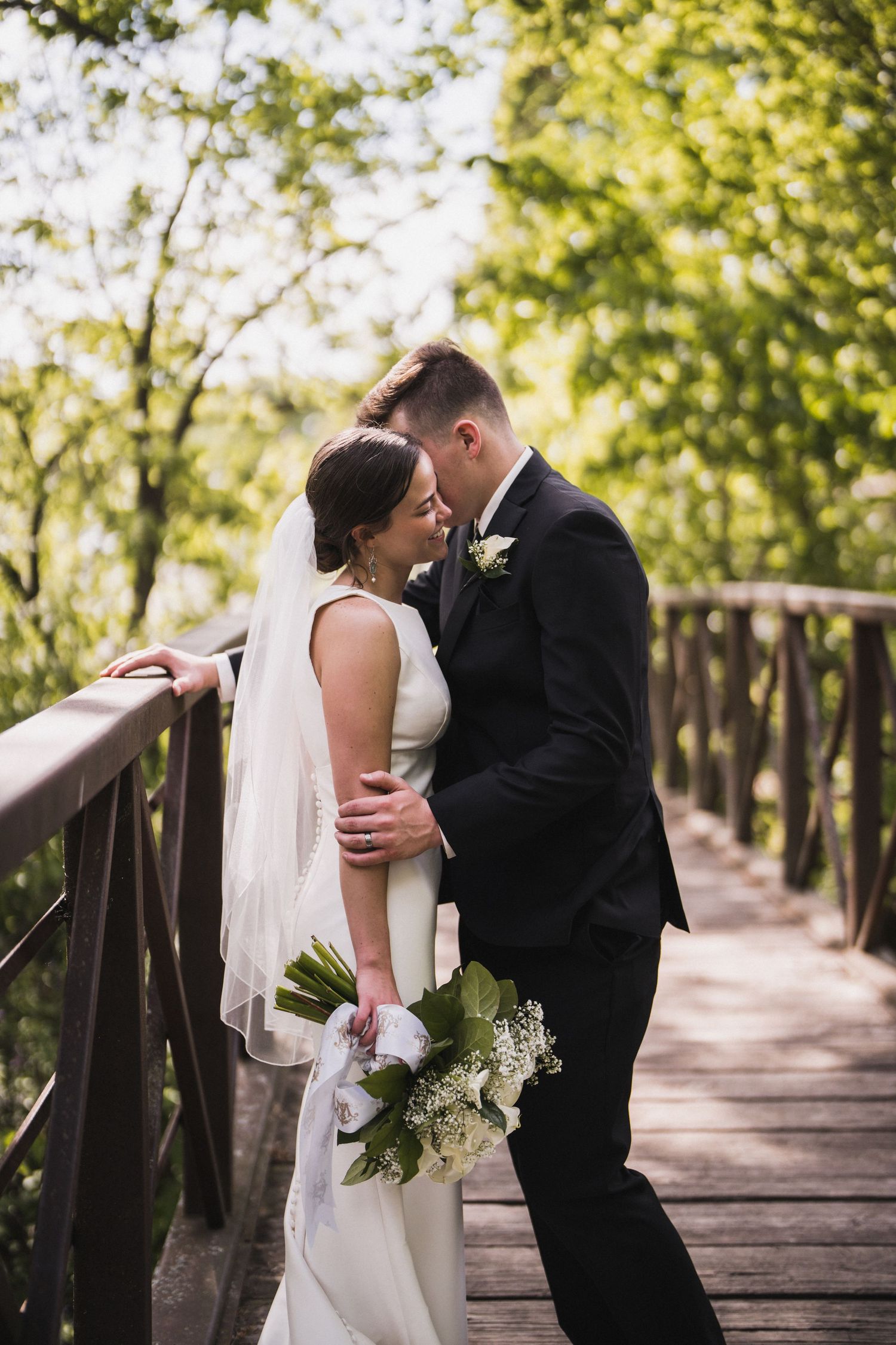 Newlyweds share intimate moment on wooden bridge surrounded by greenery.