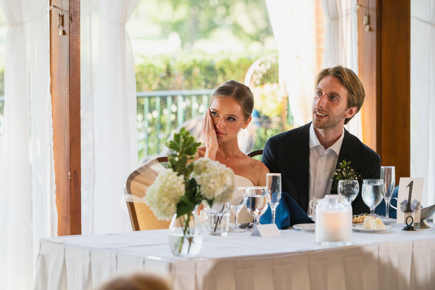 Bride and groom seated at sweetheart table during wedding reception, listening to toasts with flowers nearby.