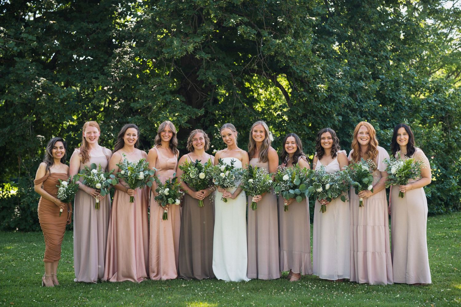 Nine bridesmaids in mismatched blush and taupe gowns holding greenery bouquets with bride in white dress.