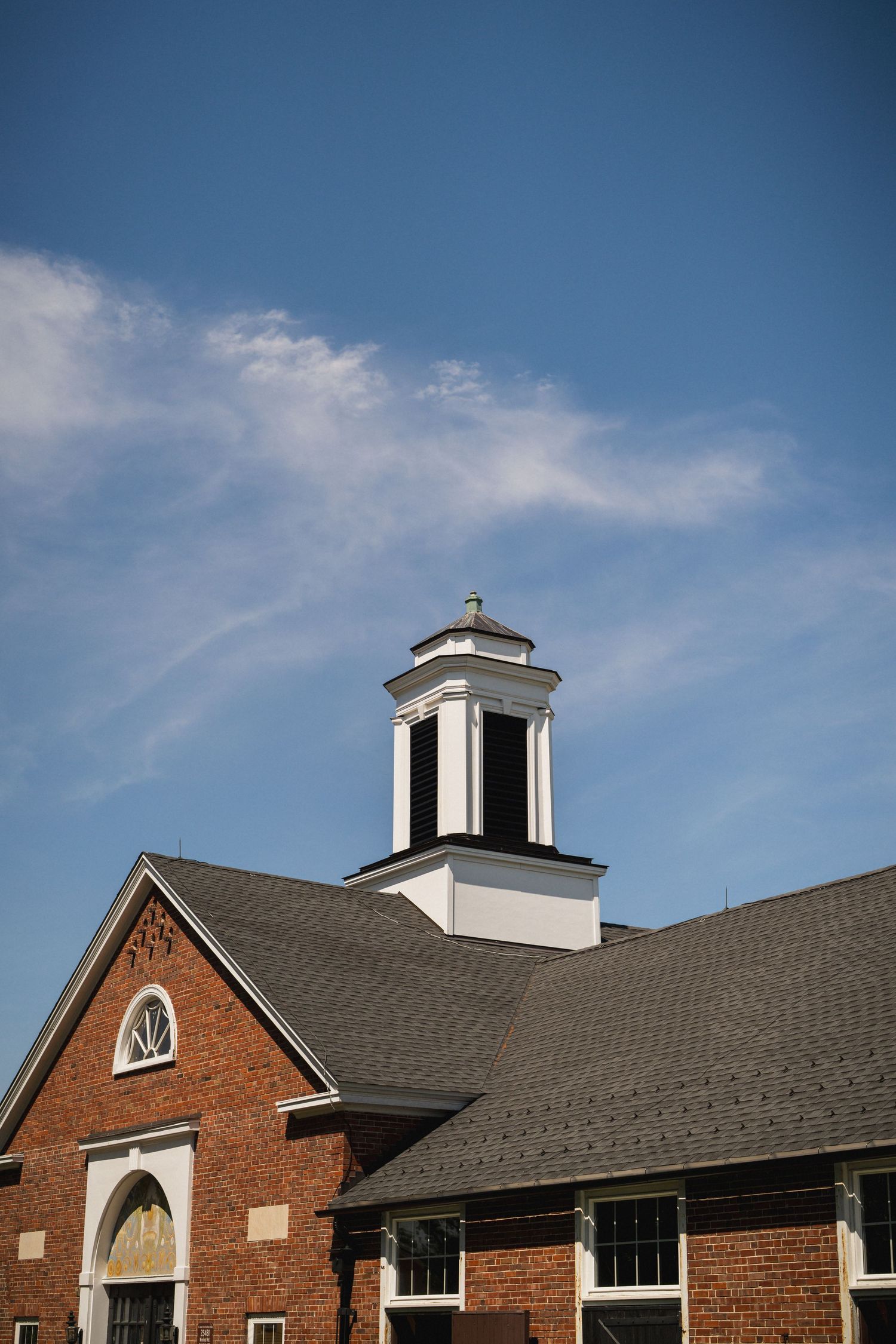 Red brick church with white cupola tower against blue sky, featuring arched windows and gray shingled roof.