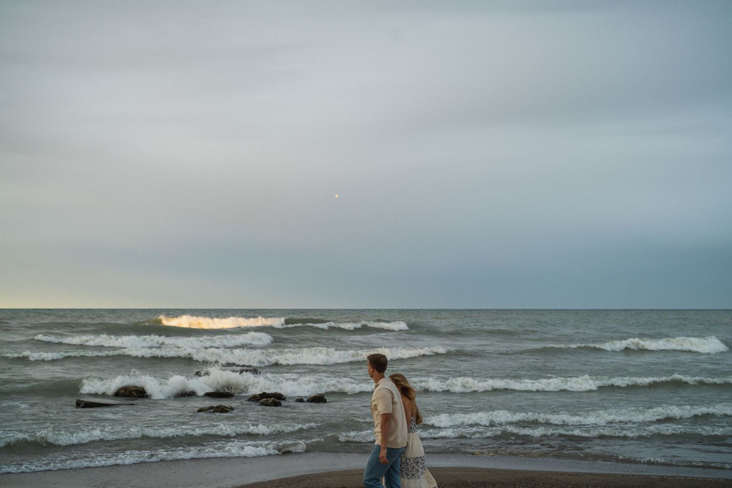 A lone person stands on a sandy beach gazing at rolling ocean waves under a cloudy gray sky at dusk.